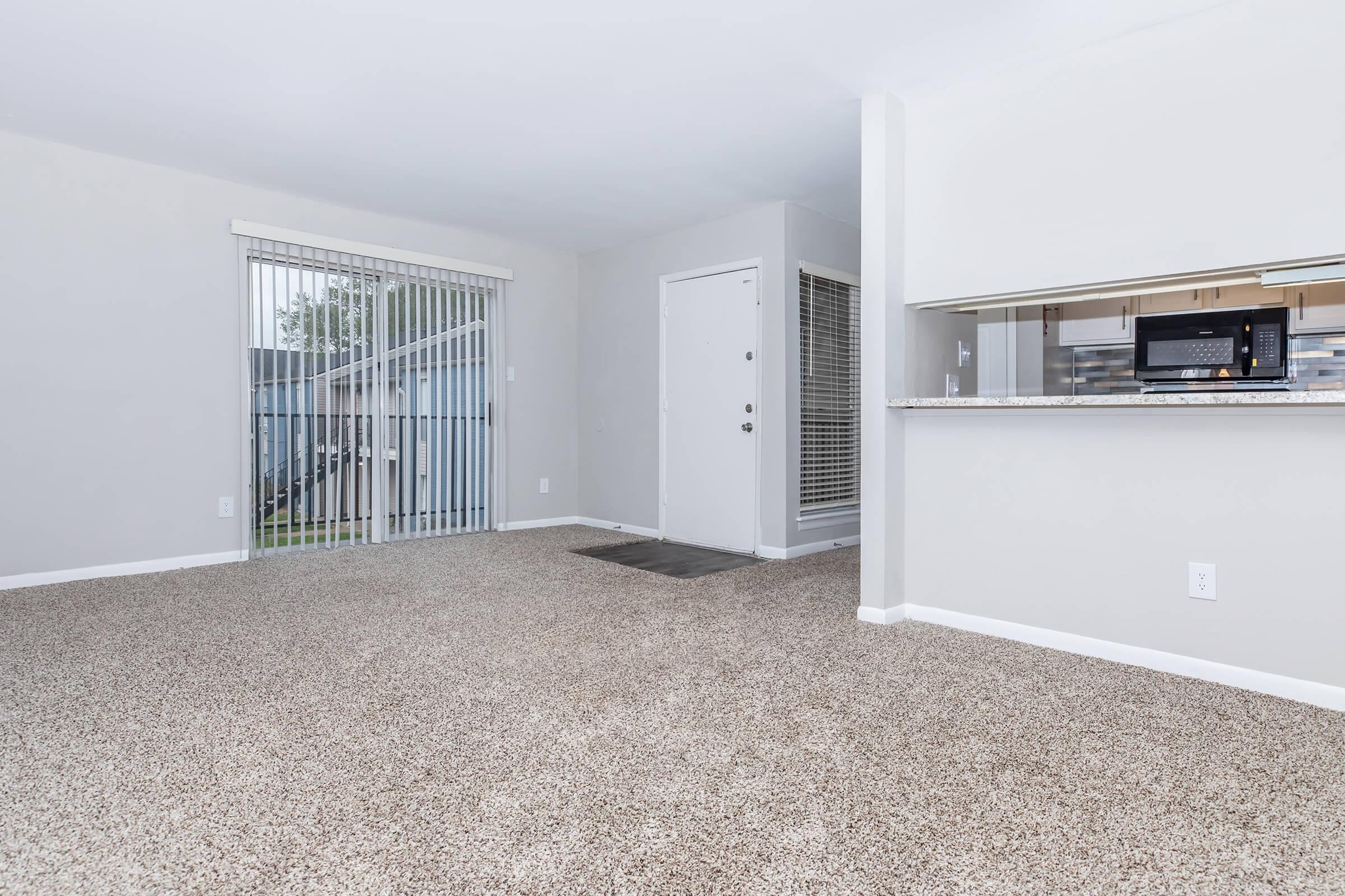 Spacious living area with light grey walls and a beige carpet. Large window with vertical blinds allowing natural light. Doorway leading to the outside and a kitchen area with a modern design visible in the background. Simple and inviting atmosphere suitable for a cozy home.