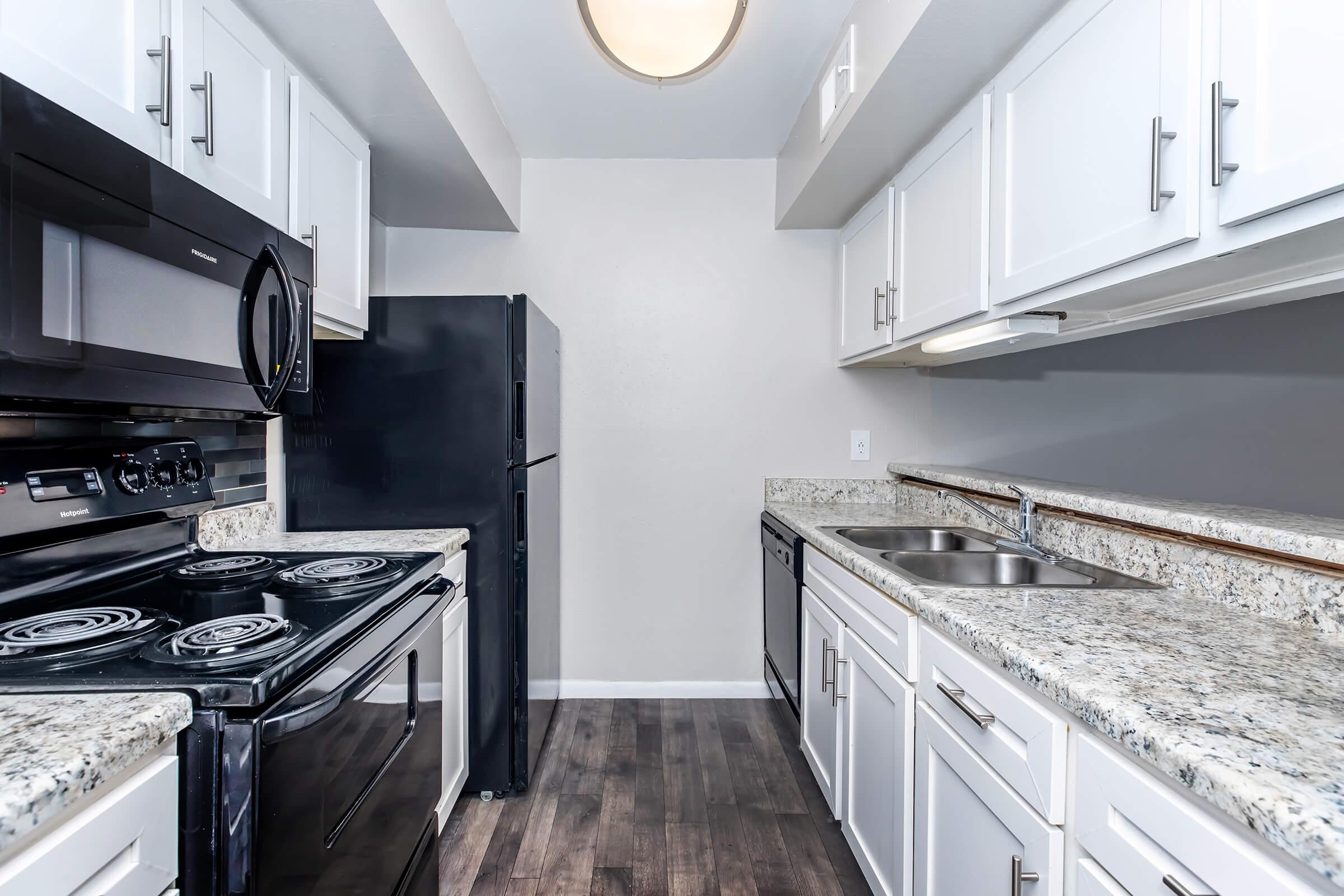 A modern kitchen featuring white cabinetry, granite countertops, a black stove and microwave, and a black refrigerator. The kitchen has double sinks and is well-lit by a ceiling fixture, with a warm color scheme and wooden flooring.
