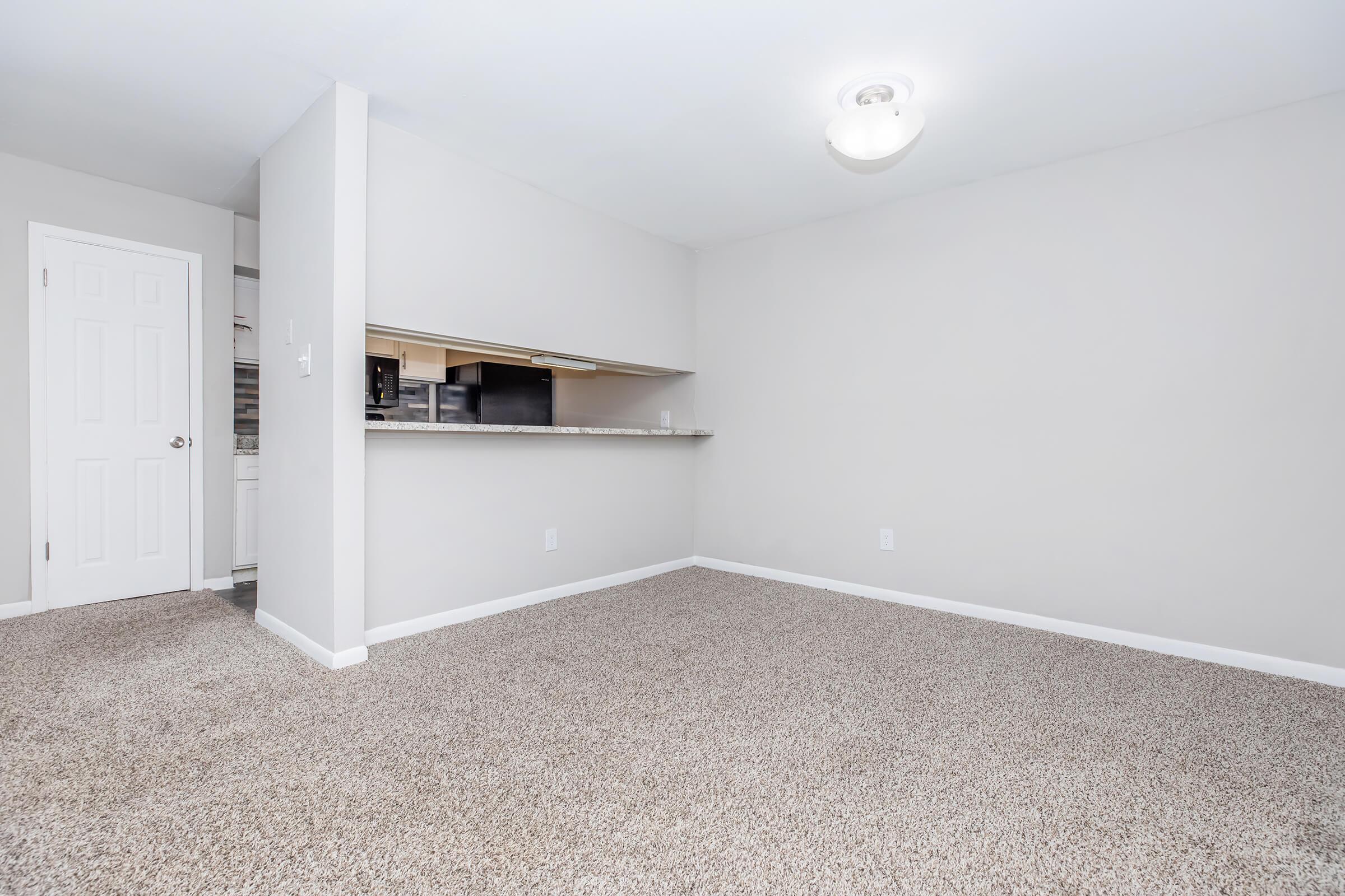 A spacious, empty room with light gray walls and carpeted flooring. There is a kitchen area visible in the background with a bar counter. A door is on the left, leading to another room, and a ceiling light fixture is present. The overall atmosphere is bright and inviting.