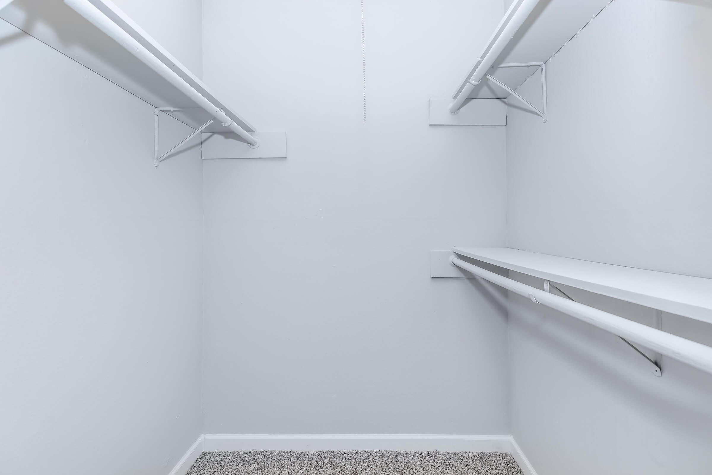 A clean, empty closet with white walls, featuring two horizontal metal rods for hanging clothes and a single shelf along one side. The floor is covered with light-colored carpet. The overall appearance is minimalistic and spacious.