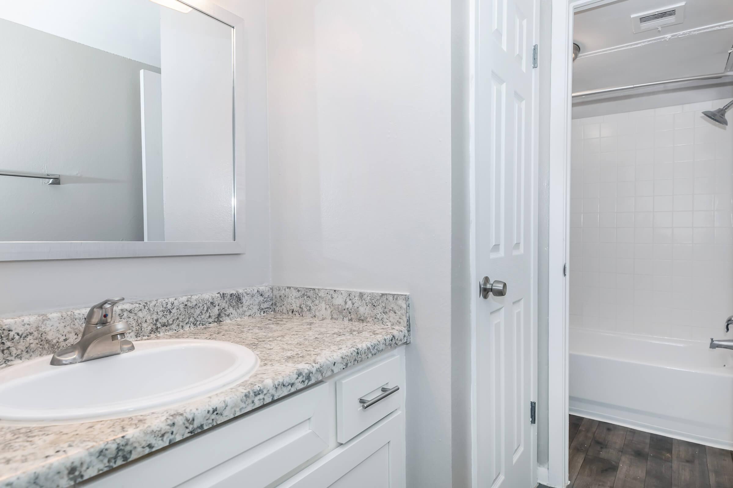 A bright, modern bathroom featuring a white sink with a granite countertop, a large mirror above the sink, and a shower/tub combination in the background. The walls are painted light, and the flooring is dark wood, creating a clean and contemporary aesthetic. A door leads to another room.