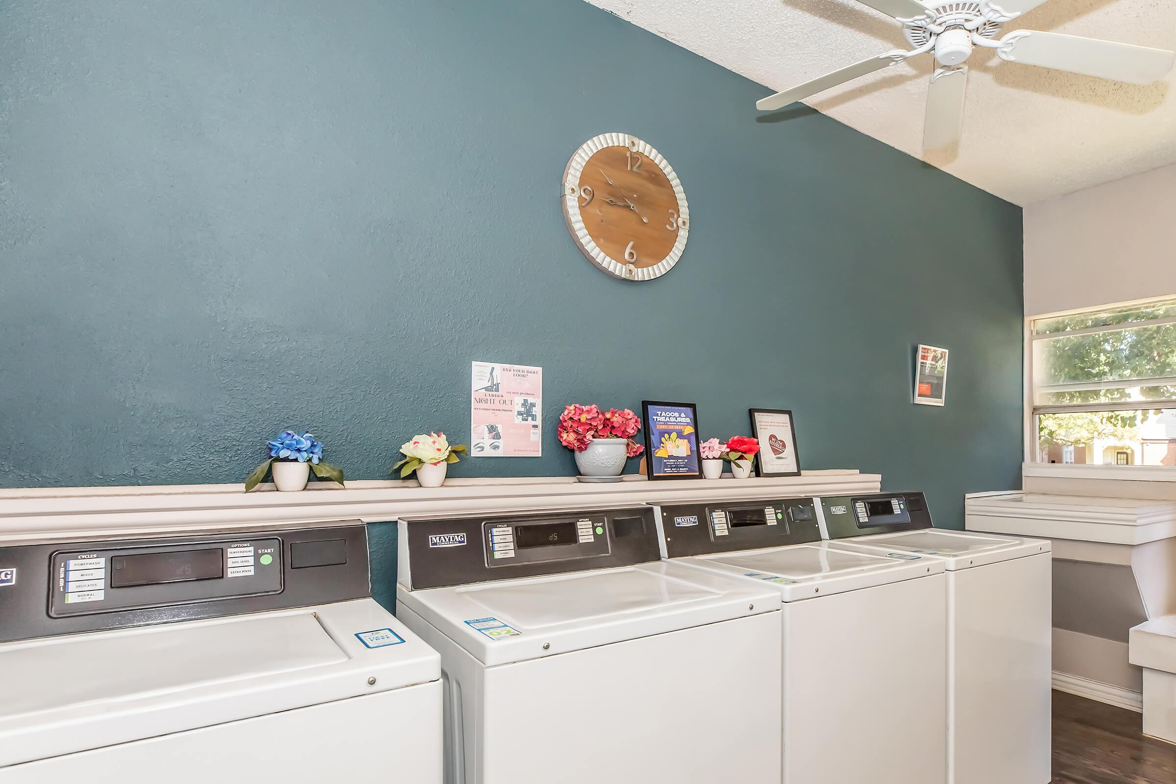 A bright laundry room featuring several white washing machines and dryers lined up against a teal wall. Decorative flower arrangements and framed pictures add a touch of color, while a clock is mounted on the wall. The space is well-lit with a ceiling fan for ventilation.