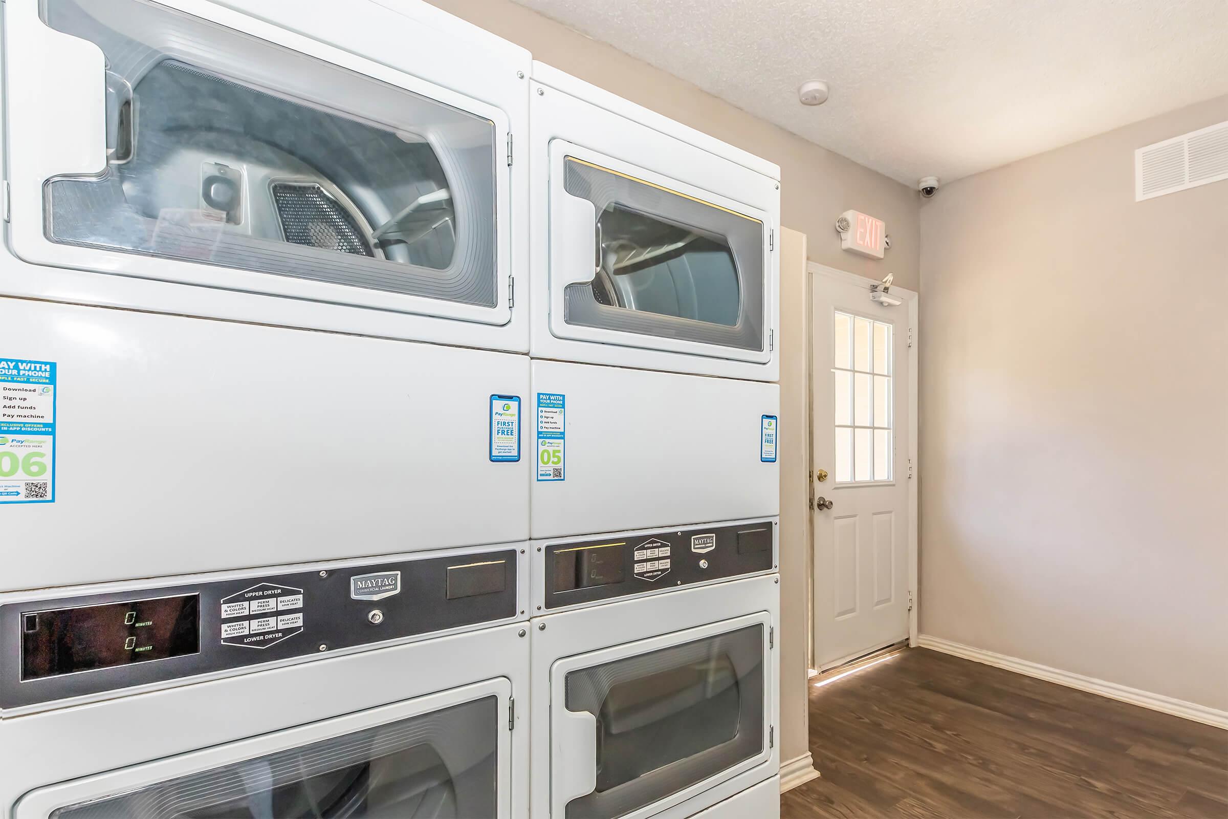 A clean, well-lit laundry room featuring stacked white washing machines and dryers. The machines have visible controls and informational stickers. There is a door leading outside, and the flooring is dark wood, creating a modern and tidy environment.