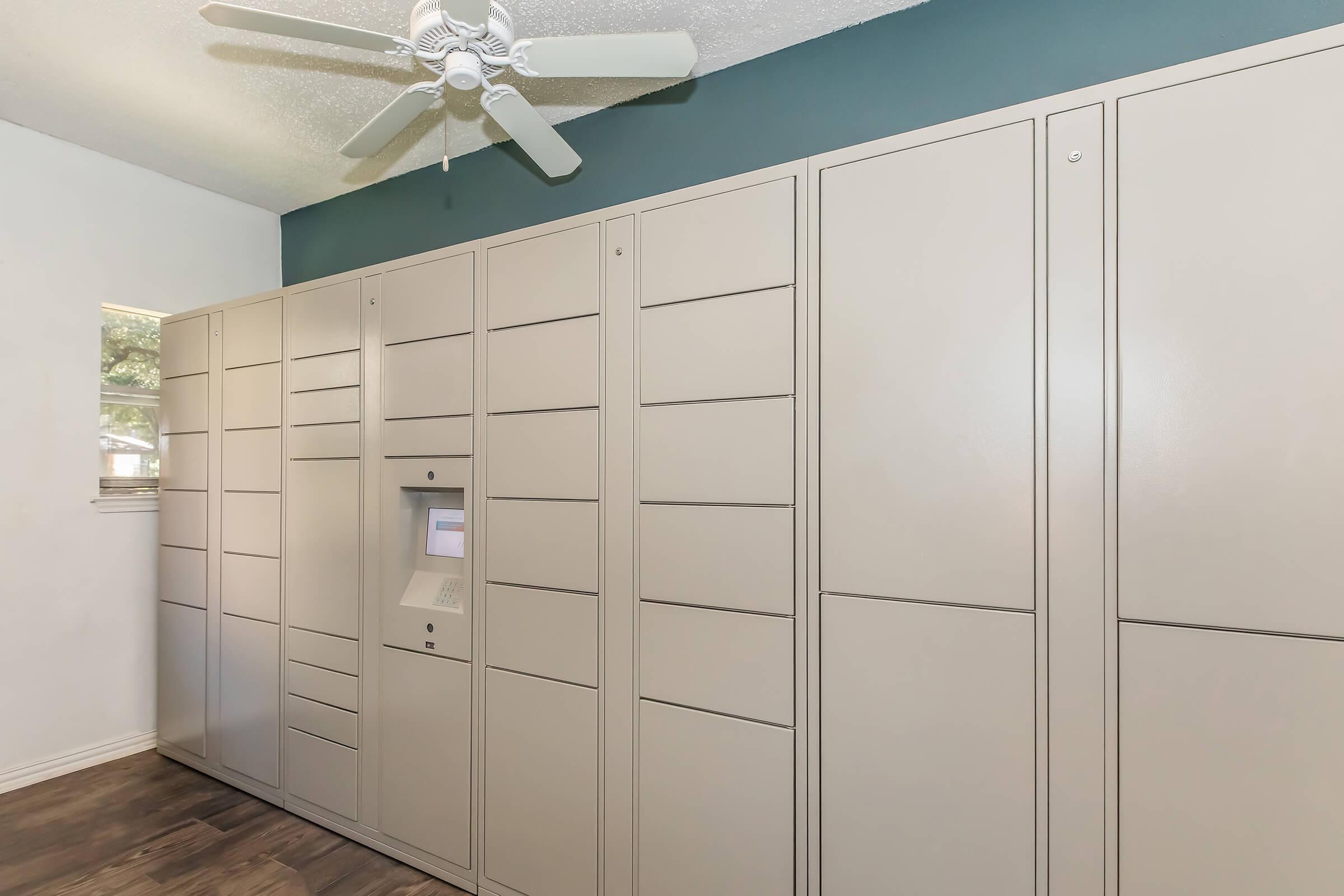 A wall-mounted package locker system featuring multiple compartments and a central control panel. The lockers are light gray, with a blue accent wall behind. A ceiling fan is visible above, and the flooring is a dark wood finish. Natural light filters in through a nearby window.