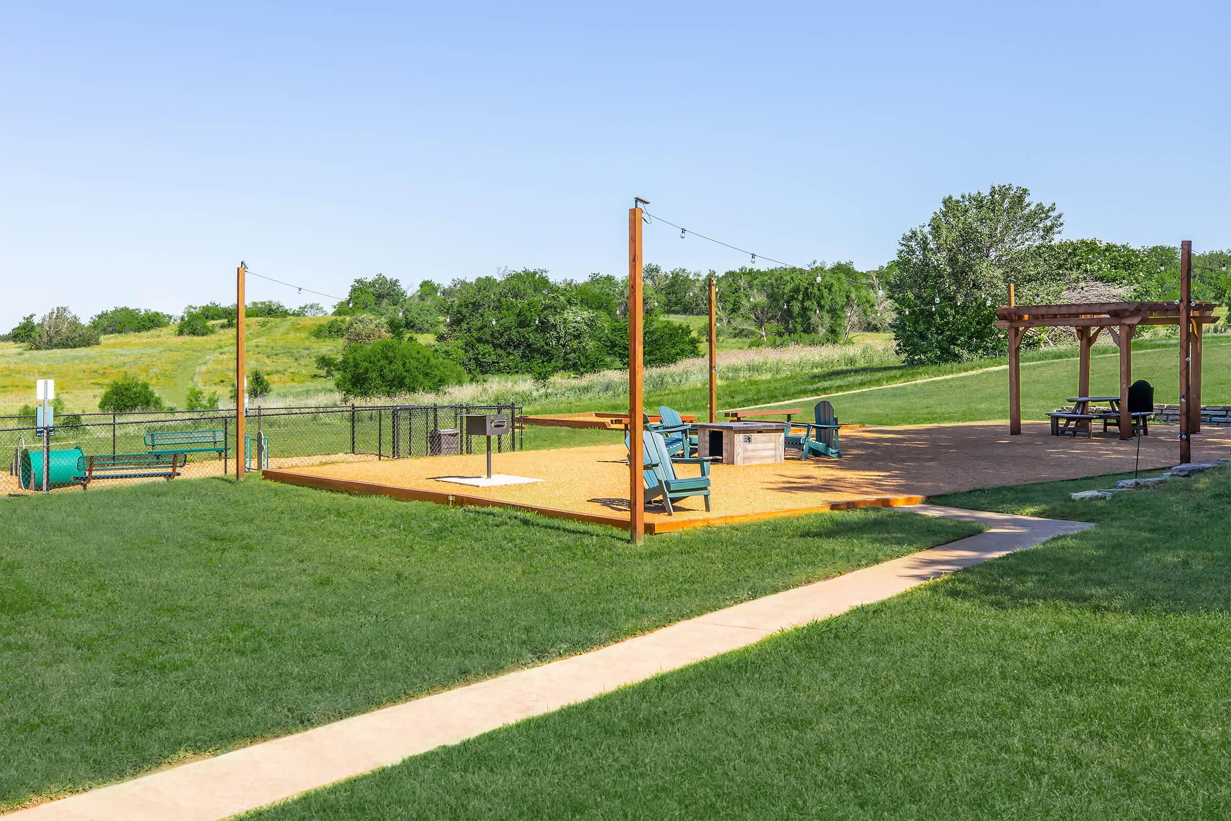A spacious outdoor area featuring green grass and a gravel path, with a seating area that includes wooden chairs and a pergola. In the background, there are trees and a fenced perimeter, providing a peaceful, recreational space for relaxation and gathering.