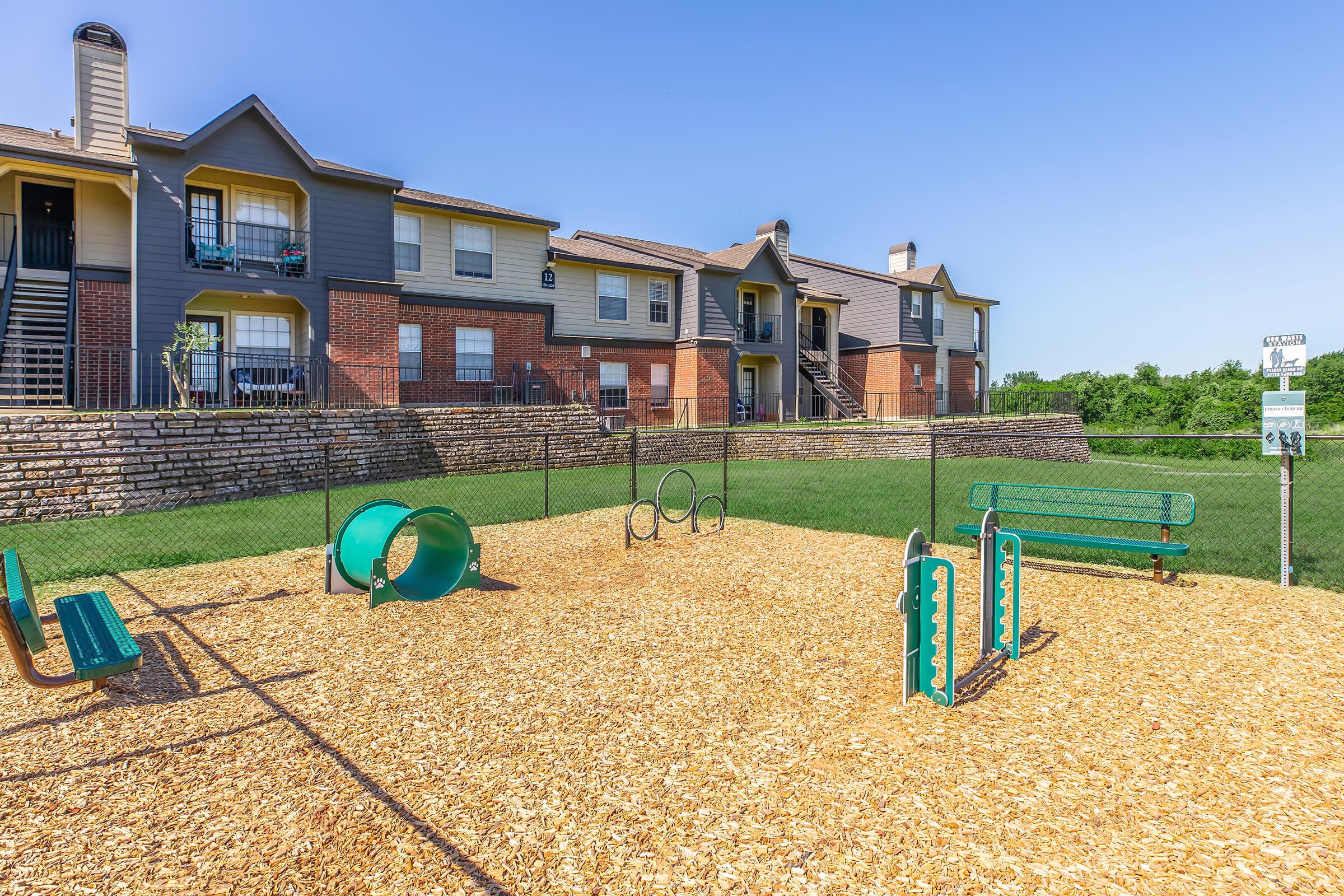 Playground area with a rubber mulch surface featuring a green slide and climbing structures, surrounded by a safety fence. In the background, two residential buildings can be seen, with balconies and staircases. The sky is clear and blue, and the grassy area adds to the outdoor setting.