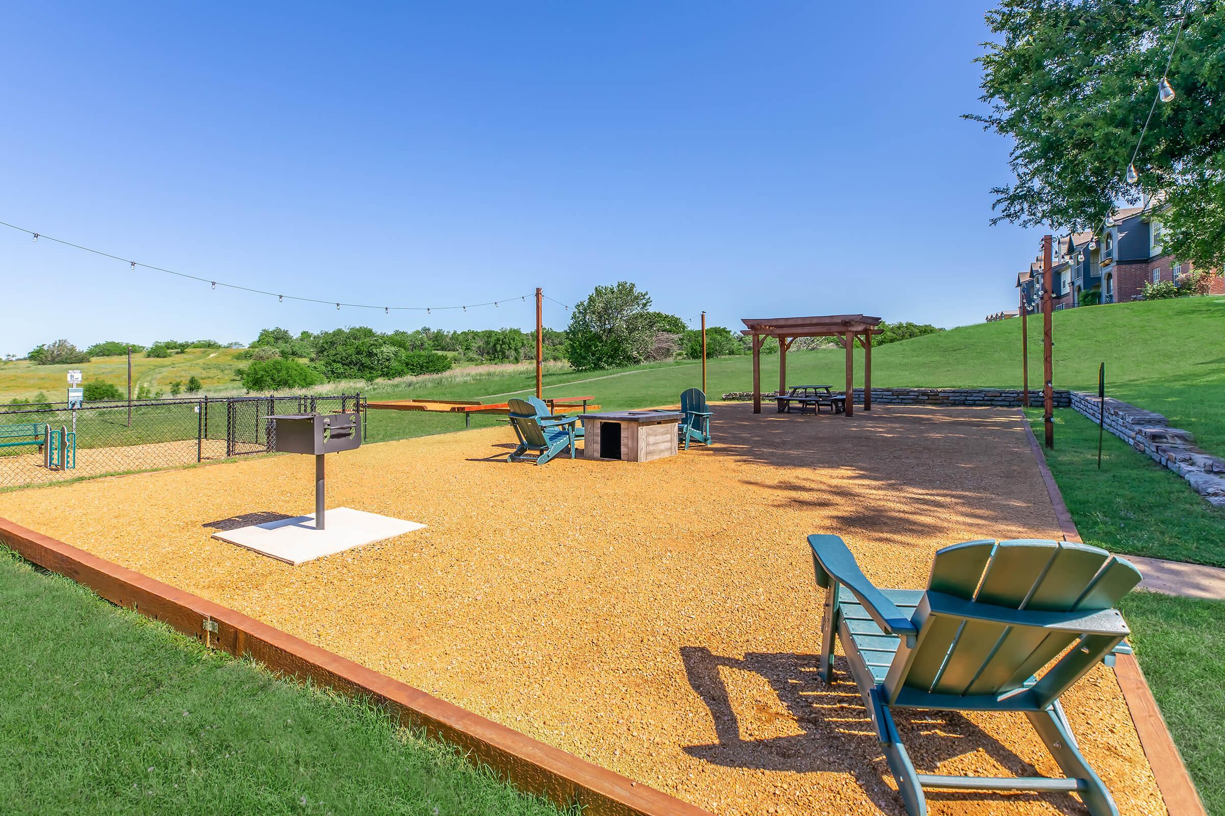 A spacious outdoor area featuring a gravel-covered ground with several green Adirondack chairs, a grilling station, and a gazebo. The background includes lush green hills under a clear blue sky, creating a relaxing atmosphere ideal for gatherings or leisure activities.