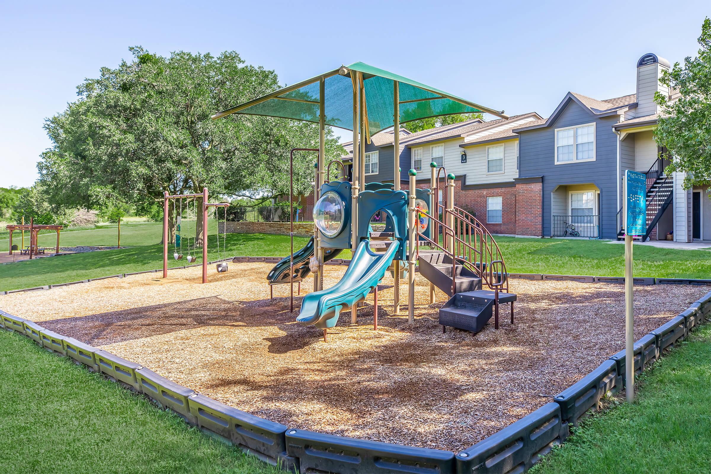 A colorful playground featuring a large play structure with slides and climbing features surrounded by a grassy area. Nearby, there are swings and a wooden fence enclosing the playground. In the background, there's a residential building with trees and blue sky above.