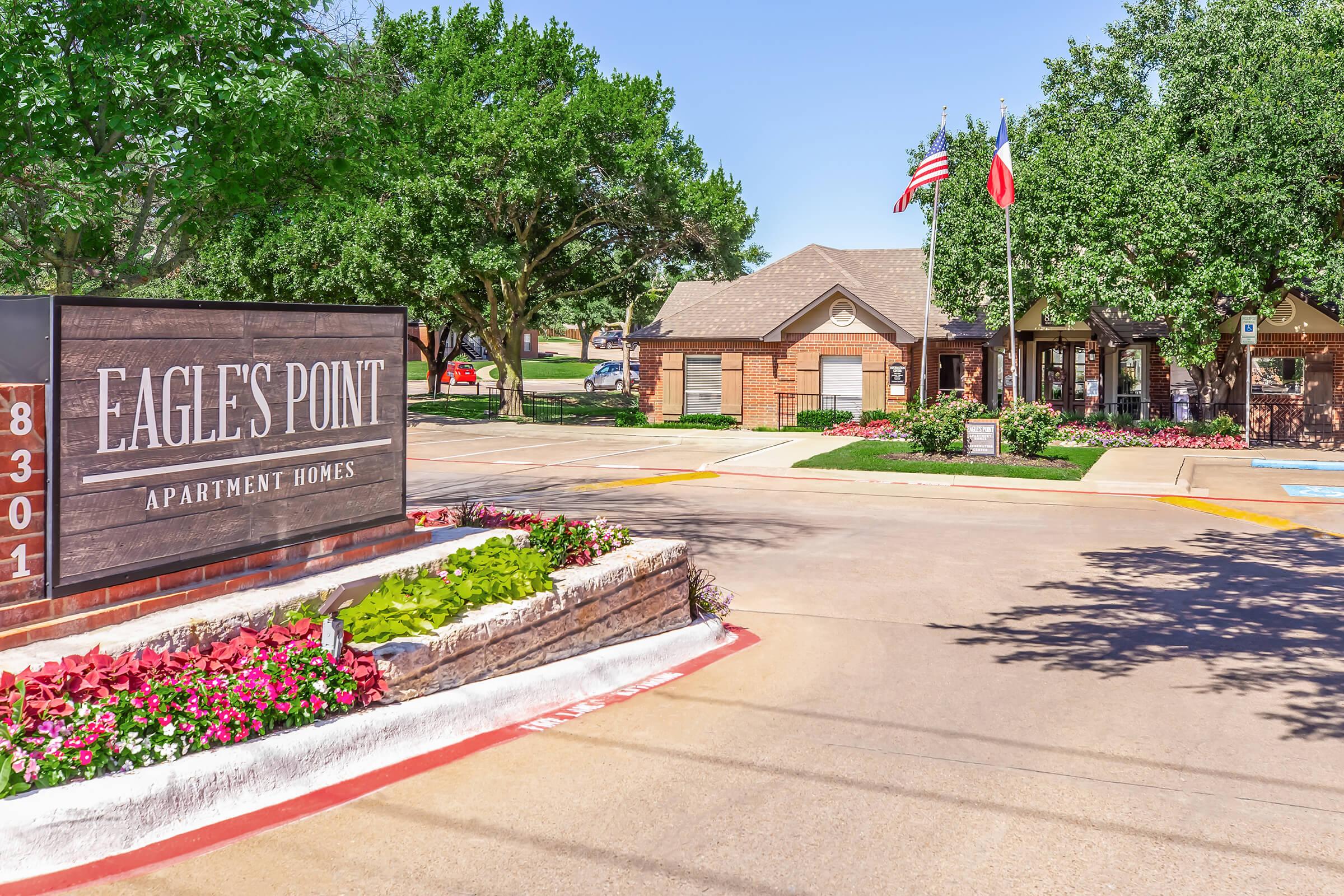 Sign for Eagle's Point Apartment Homes at the entrance, featuring lush landscaping with colorful flowers and trees. The background includes a welcoming building with flags flying, capturing a sunny day at the apartment complex.