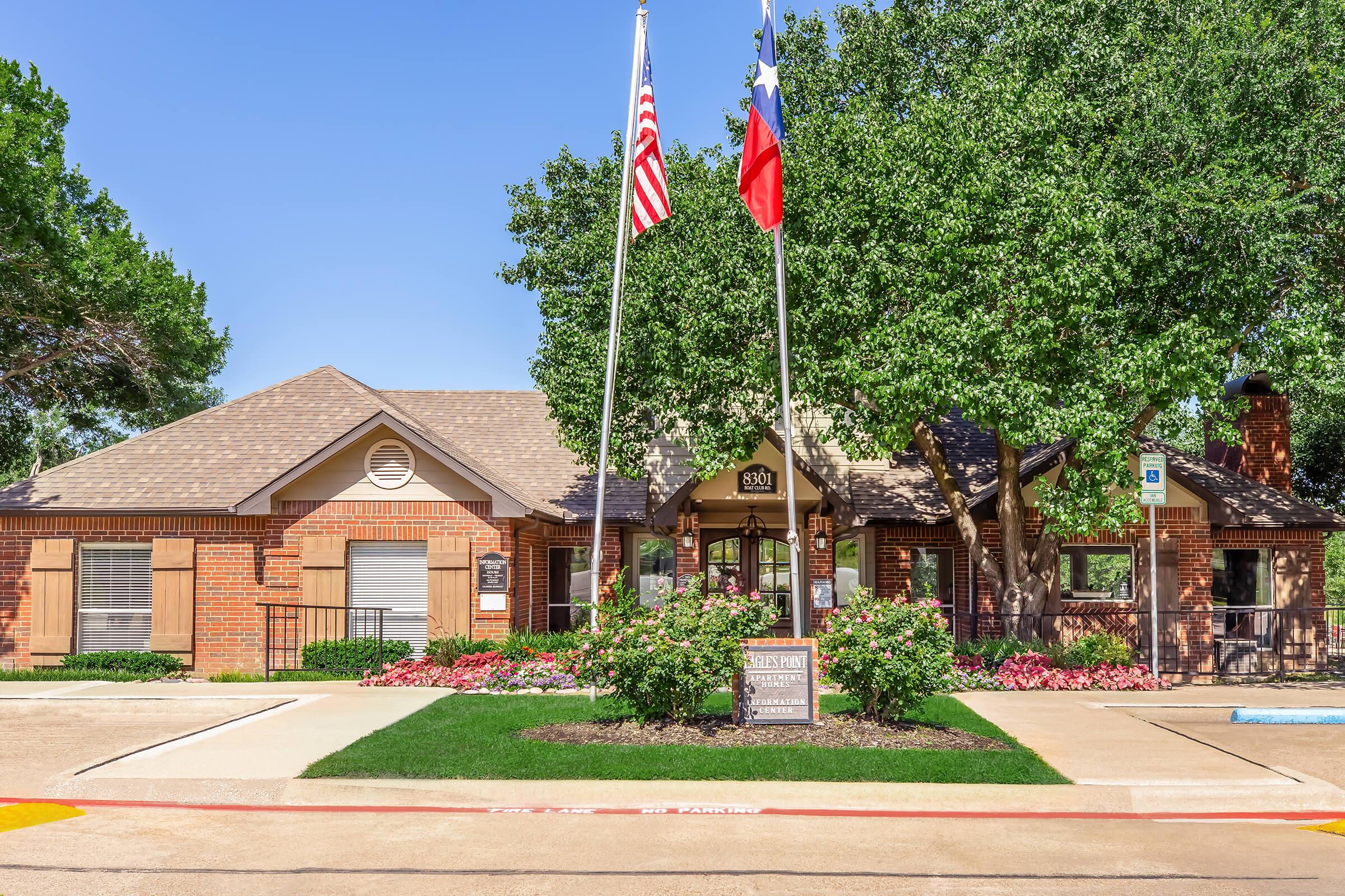 Single-story brick building with a peaked roof, surrounded by well-maintained landscaping. Two flags (U.S. and Texas) are prominently displayed on poles in front. The entrance features a sign and is framed by flowerbeds with colorful blooms. Clear blue sky overhead. Parking area visible in the foreground.