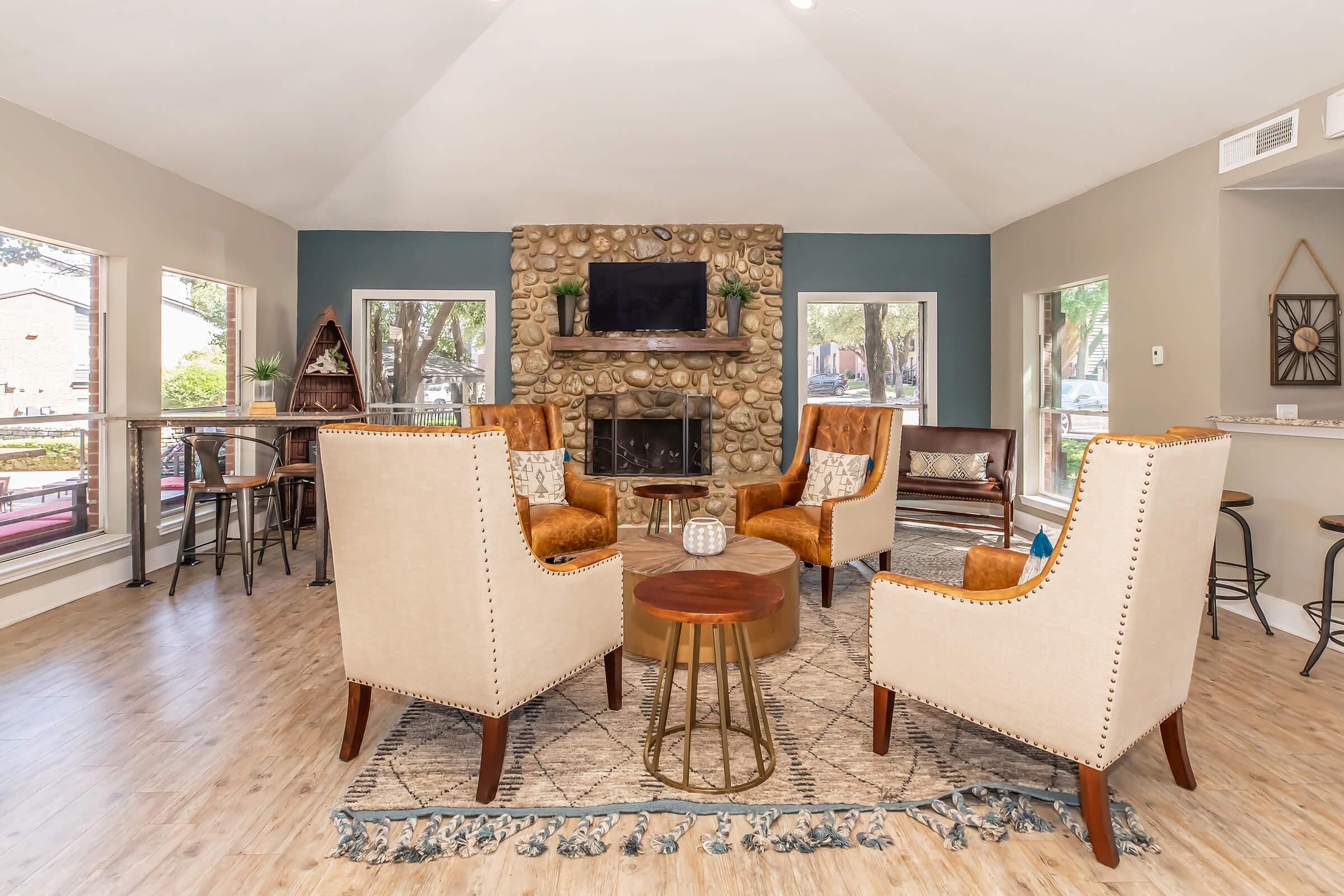 Cozy lounge area featuring two beige armchairs and orange accent chairs around a wooden coffee table. A stone fireplace is the focal point, with a TV mounted above. Large windows provide natural light, and there's a bar area with stools and a patterned rug on the floor.