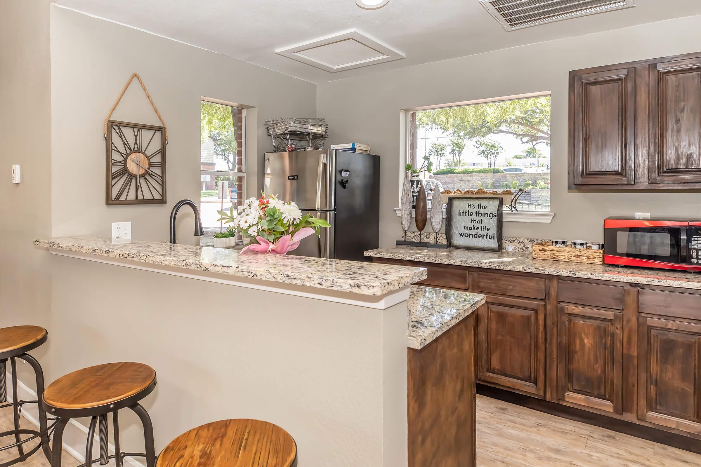 A modern kitchen featuring granite countertops, wooden cabinets, and bar stools. There is a stainless steel refrigerator, a microwave, and decorative items including a wall clock and a sign with an inspirational quote. Natural light streams in through two windows, illuminating the space.