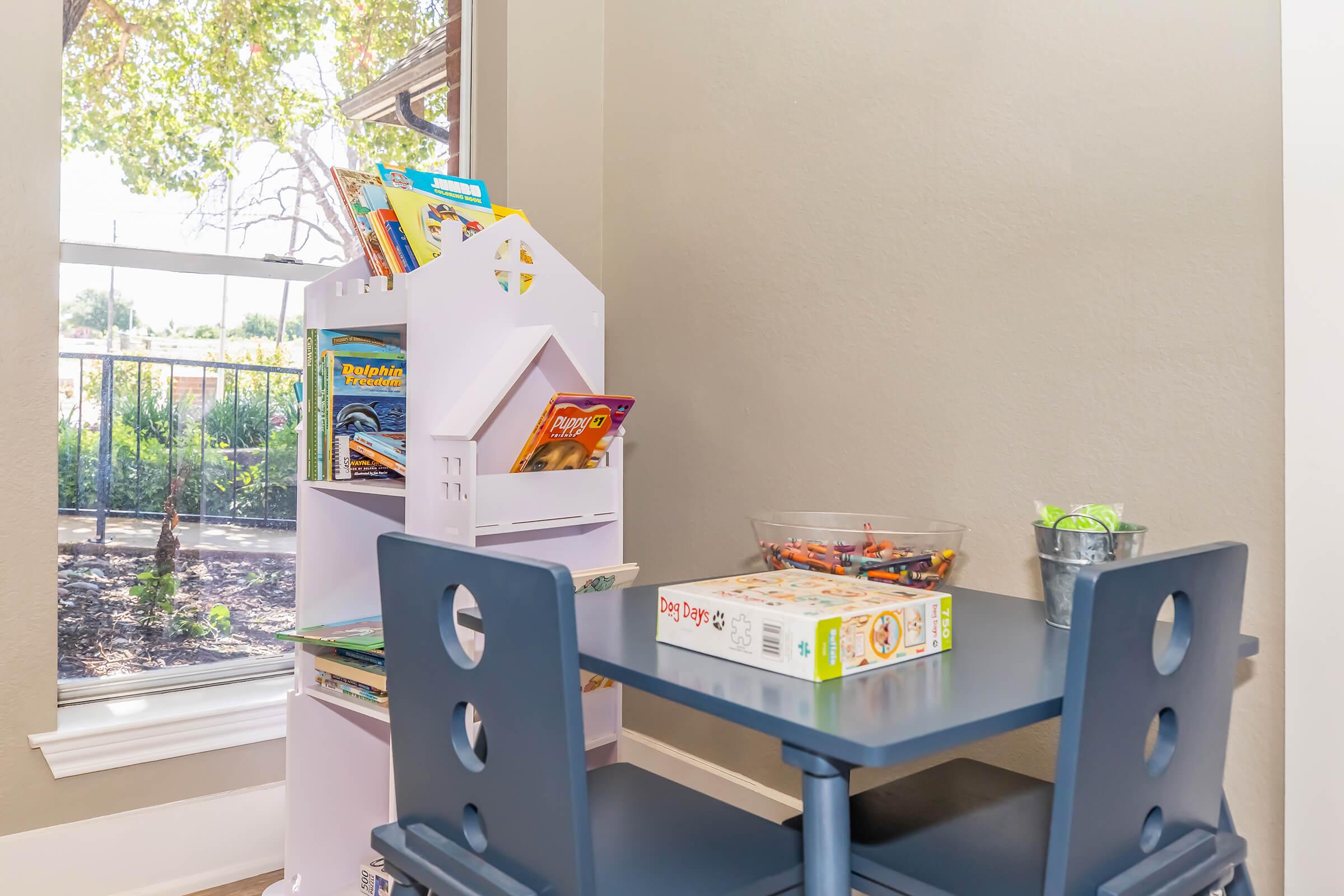 A small children's table set next to a window, featuring a stack of colorful books and board games on a shelf. A container of crayons is on the table, along with a small metal bucket. Natural light fills the space, creating a bright and inviting atmosphere for play and reading.