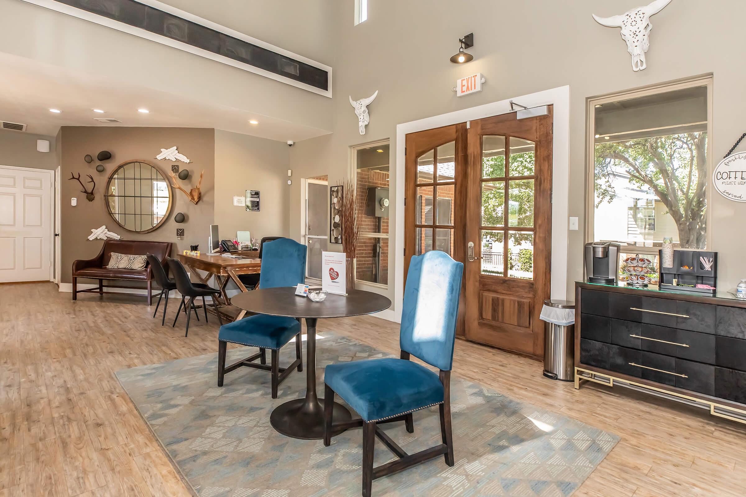 A cozy interior space featuring a small round table with two blue chairs, a welcoming reception area with a sofa, decorative wall art, and a large mirror. Natural light streams through glass doors, and a small coffee station is visible in the corner.