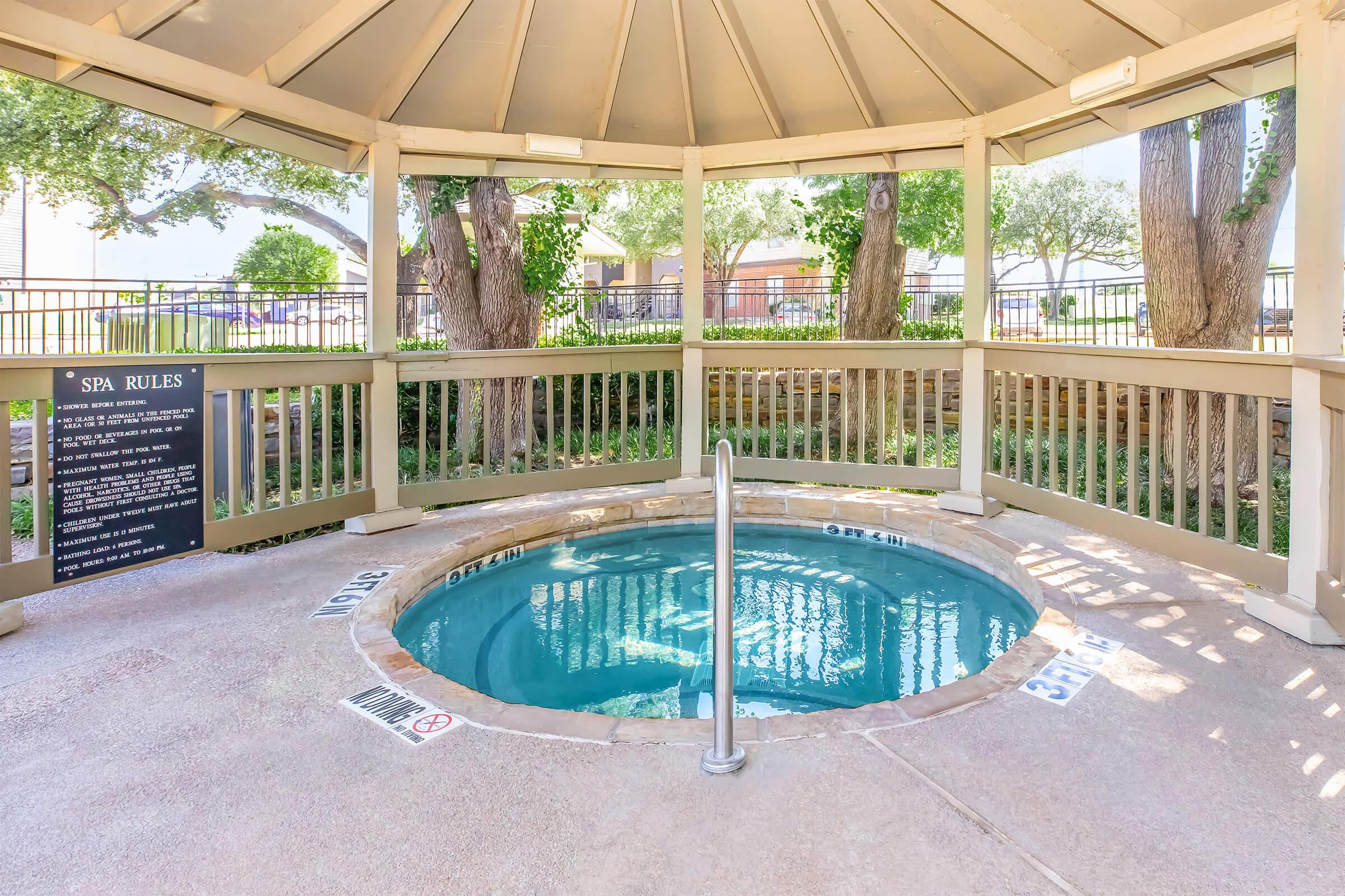 A small round hot tub located under a gazebo, surrounded by a wooden railing and trees. A sign displaying "Spa Rules" is visible nearby, along with safety information. The water is clear, and the area is well-maintained and inviting.