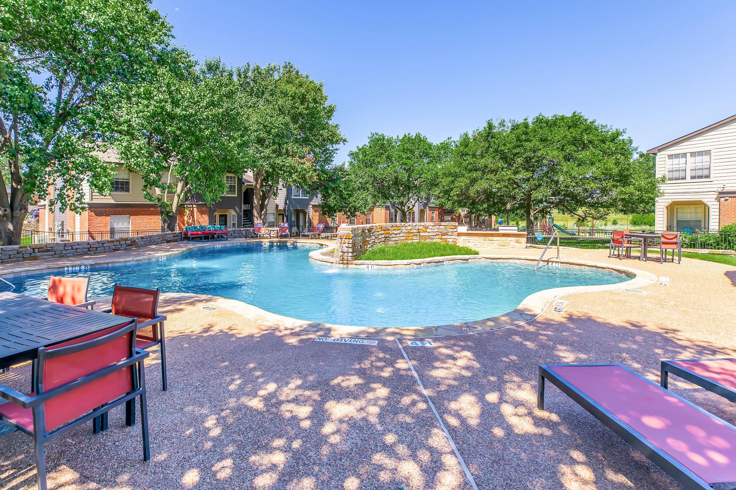 A sunny poolside area featuring a curved swimming pool surrounded by landscaped greenery. There are lounge chairs and tables with chairs positioned nearby, creating a relaxing atmosphere in a residential setting. The sky is clear, enhancing the inviting ambiance of the area.