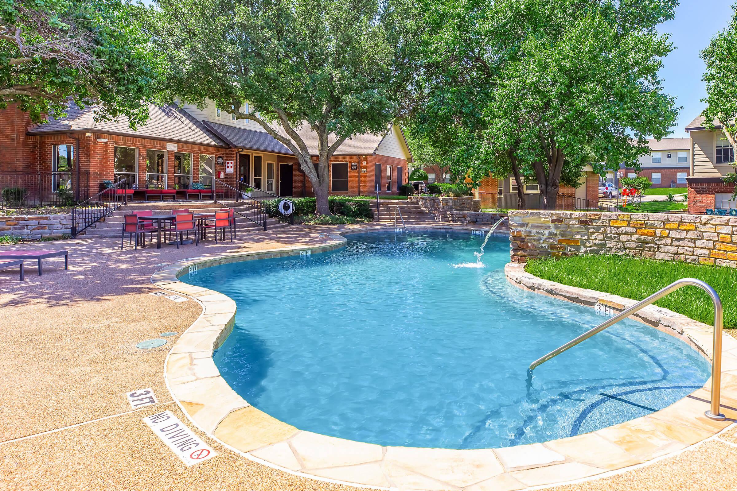 A well-maintained outdoor swimming pool, surrounded by lush green trees and manicured landscaping. The pool features a gradual entry with a handrail, a built-in water fountain, and is adjacent to a cozy seating area with red chairs. A nearby building provides amenities such as restrooms and shaded areas.