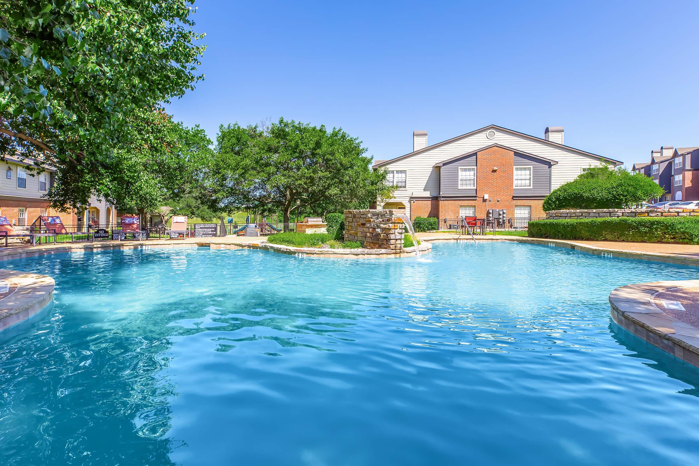 A tranquil swimming pool surrounded by lush trees and well-maintained landscaping, with a brick building in the background. Clear blue skies reflect on the water's surface, creating a serene atmosphere for relaxation. Lounge chairs are visible around the pool area, inviting enjoyment and leisure.