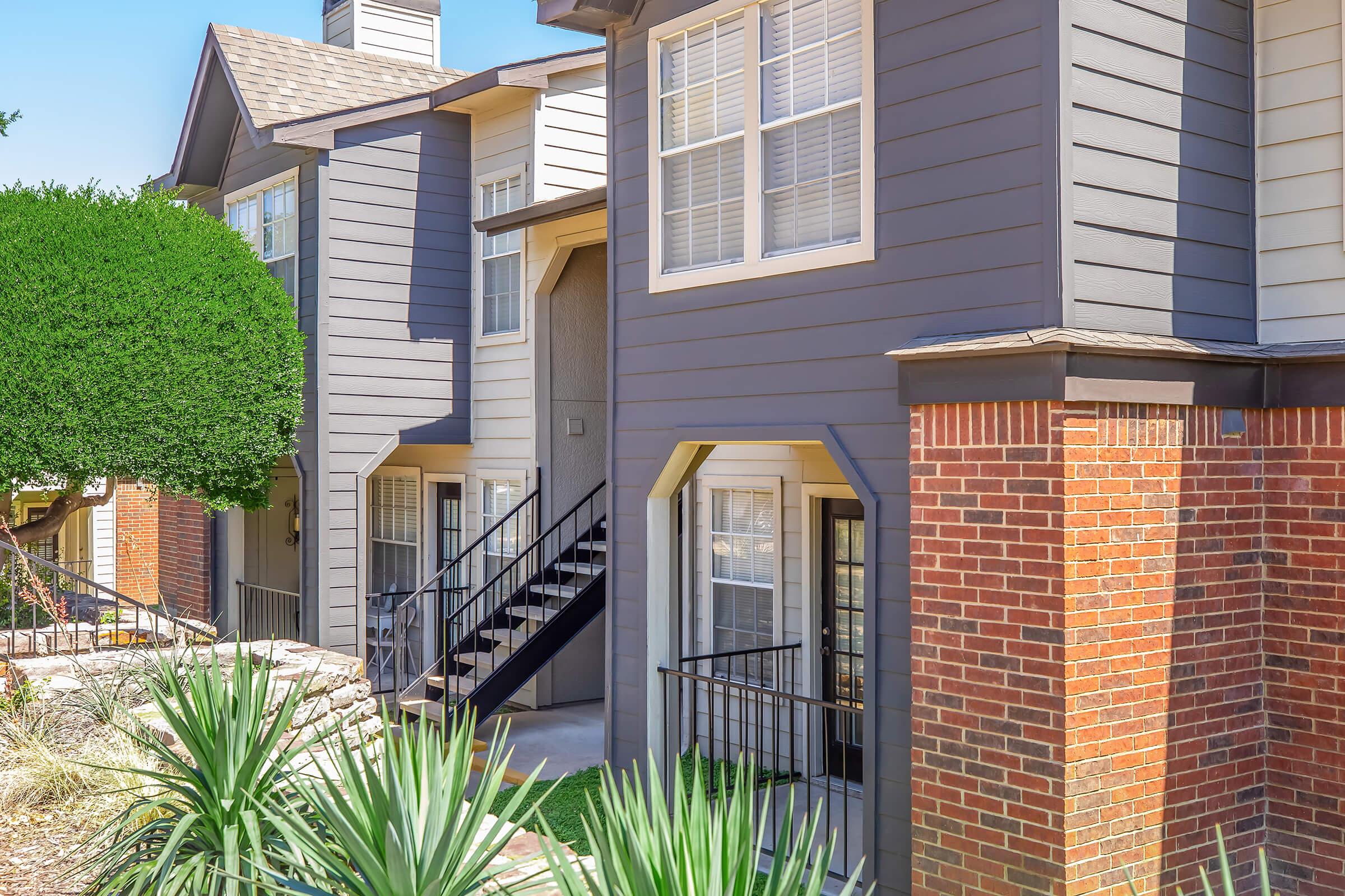 Two-story apartment building featuring a mix of gray siding and brick exterior, with green bushes and landscaping in front. A staircase leads to the second floor, and multiple windows provide natural light. The setting appears well-maintained and inviting.