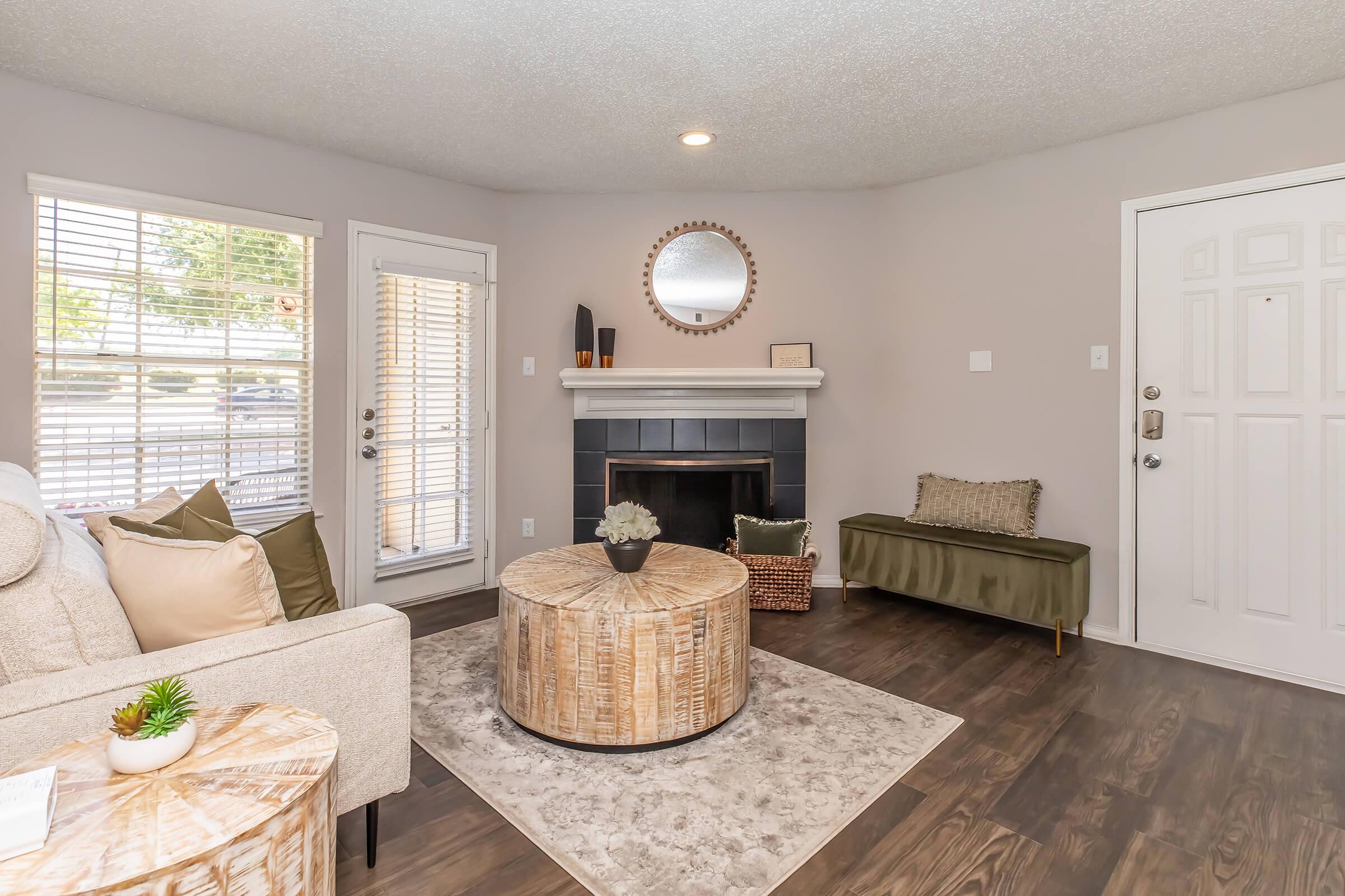 Cozy living room featuring a neutral color scheme, a textured area rug, a round coffee table, and a modern fireplace. Natural light streams through large windows with blinds. A circular mirror hangs above the fireplace, complemented by decorative elements on the mantel and seating area.