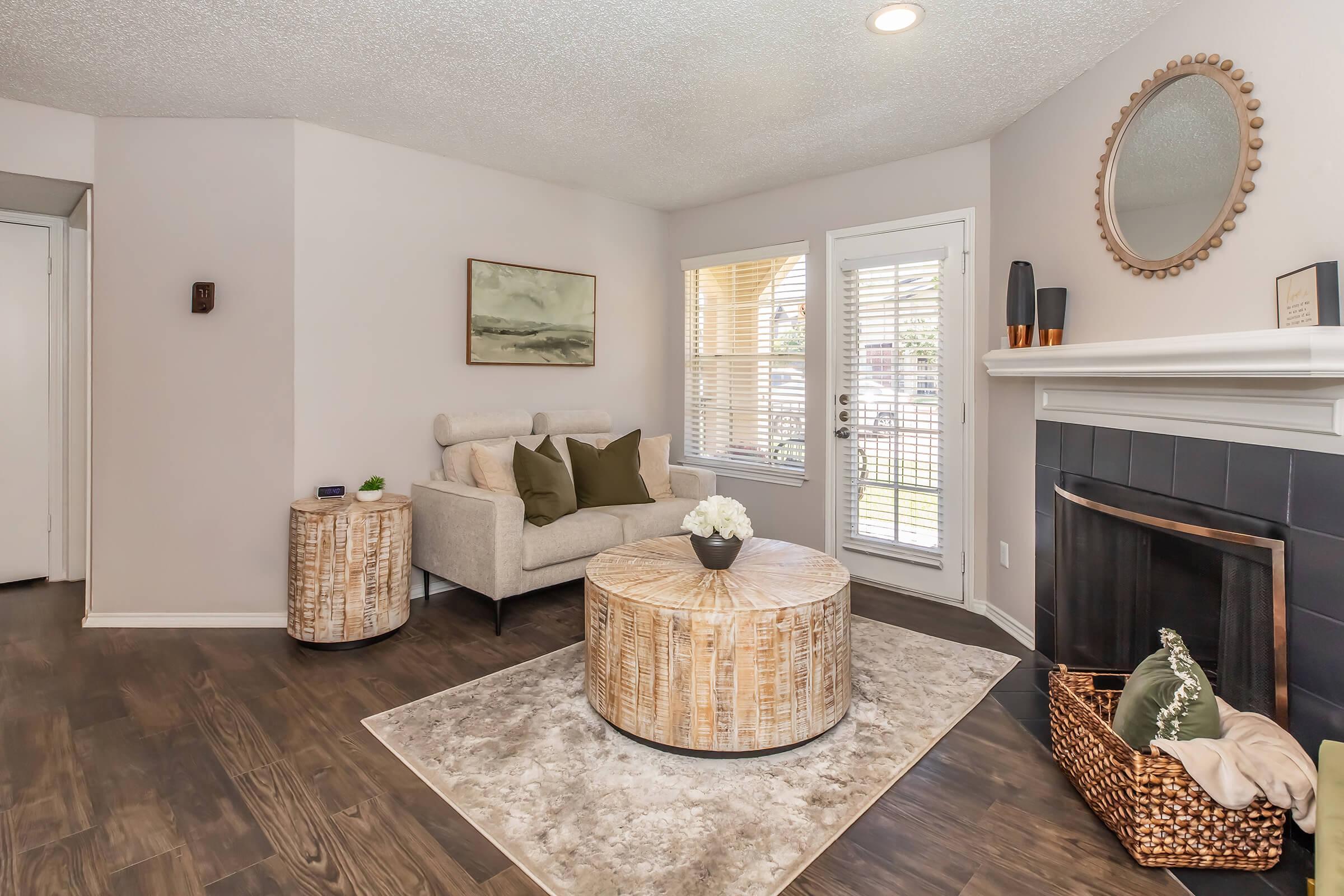 Cozy living room featuring a light-colored sofa with green cushions, a round coffee table, and a large area rug. There’s a fireplace with decorative items, a mirror on the wall, and large windows with blinds allowing natural light. Two accent tables are placed beside the sofa.