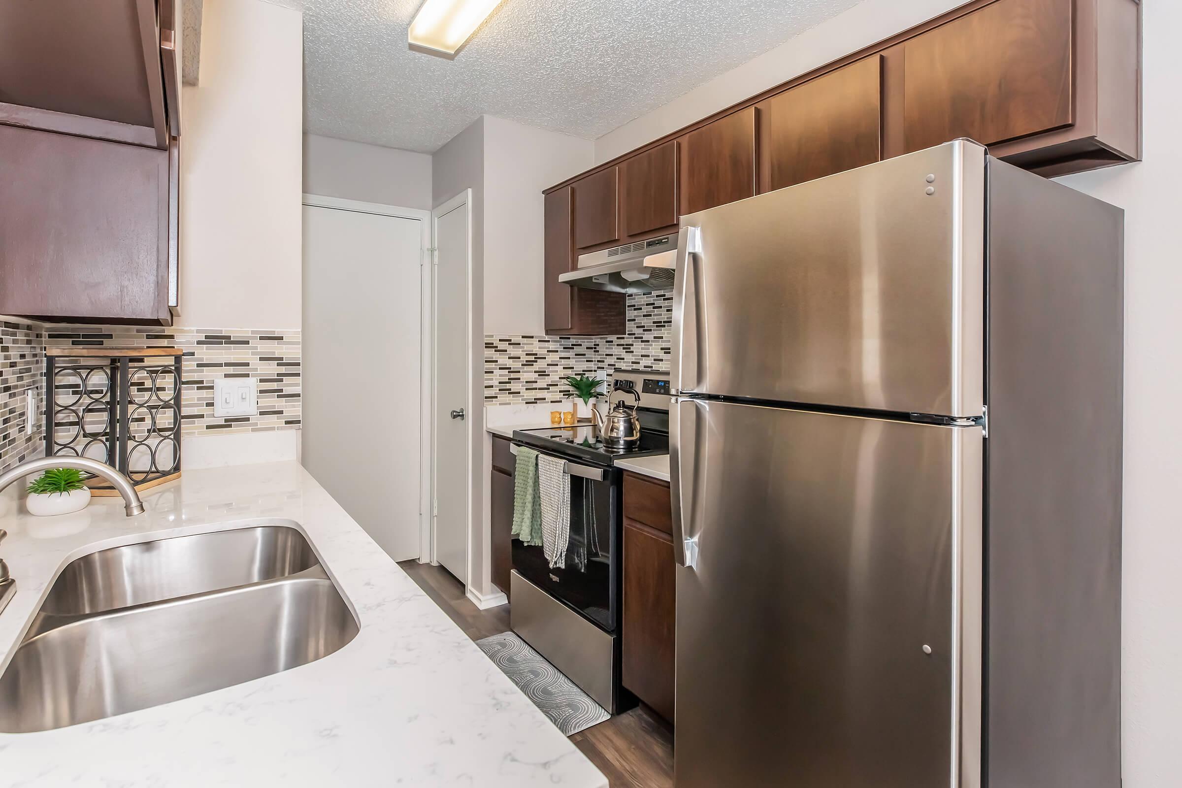 Modern kitchen featuring dark wood cabinetry, stainless steel appliances including a refrigerator and an oven, and a marble countertop. A small sink is visible, along with mosaic tile backsplash and decorative elements on the counter. The kitchen has a contemporary design with ample lighting.