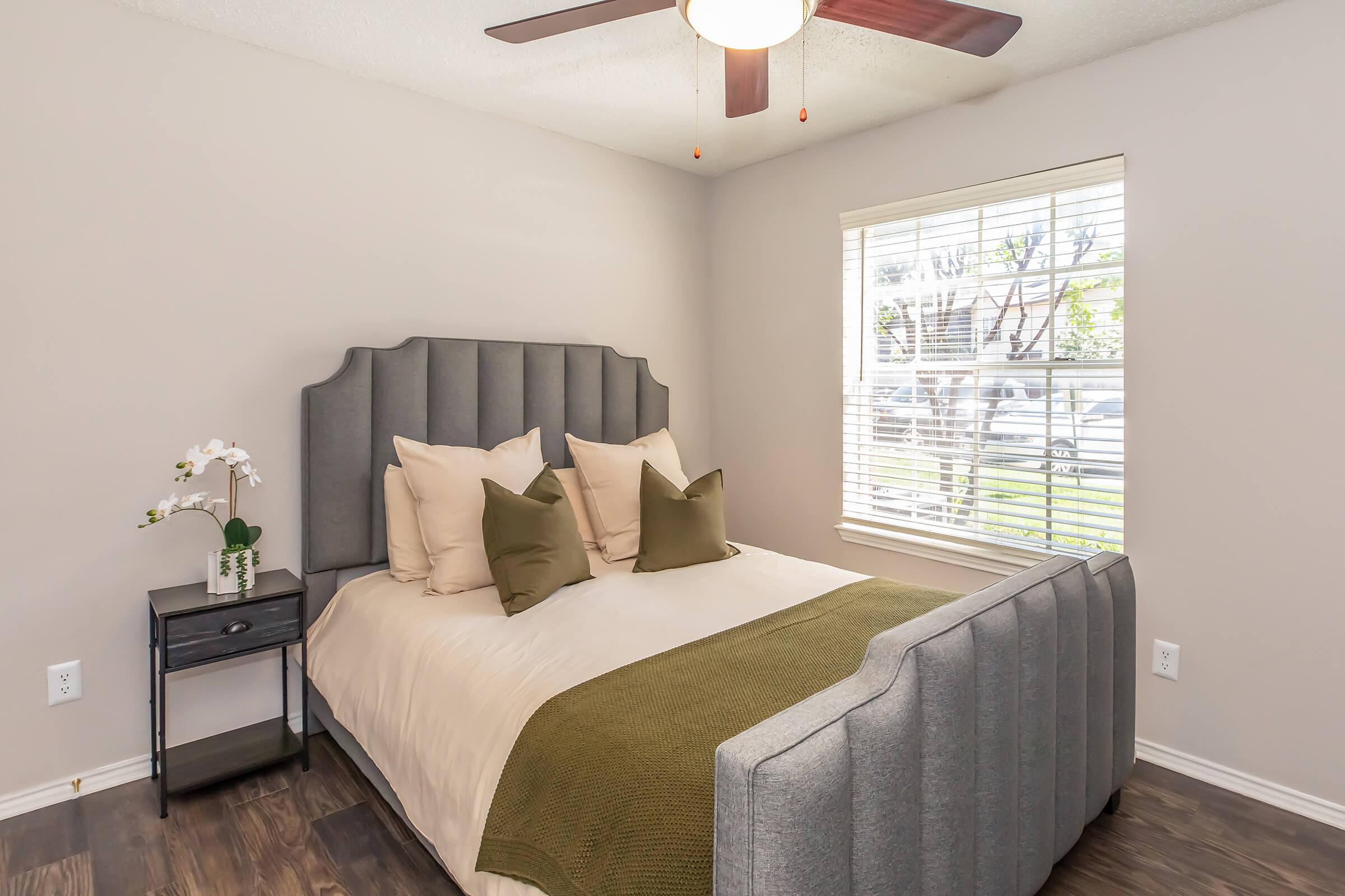 A cozy bedroom featuring a gray upholstered bed with beige and green decorative pillows. A nightstand with a vase of white flowers sits beside the bed. Natural light filters through a window with white blinds, illuminating the space with a neutral color palette and wooden flooring.
