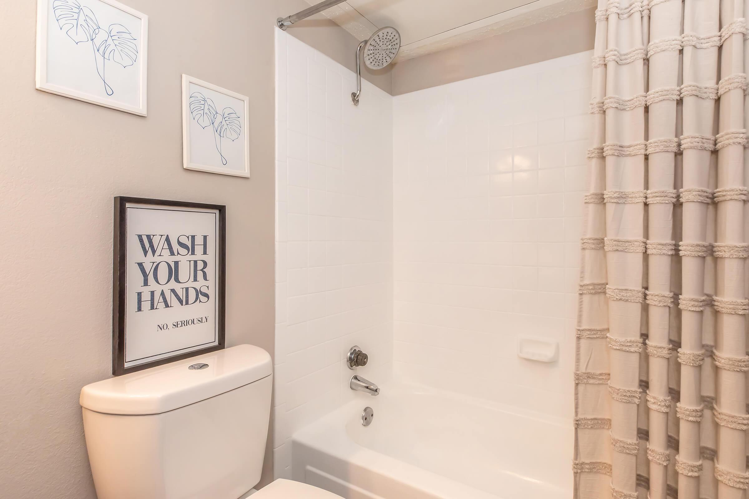 A clean and modern bathroom with a white bathtub and shower. The wall features a decorative sign that reads "WASH YOUR HANDS" and two framed botanical prints. The shower area is enclosed with a clear glass door, and a textured shower curtain adds a stylish touch. The overall color scheme is neutral and inviting.