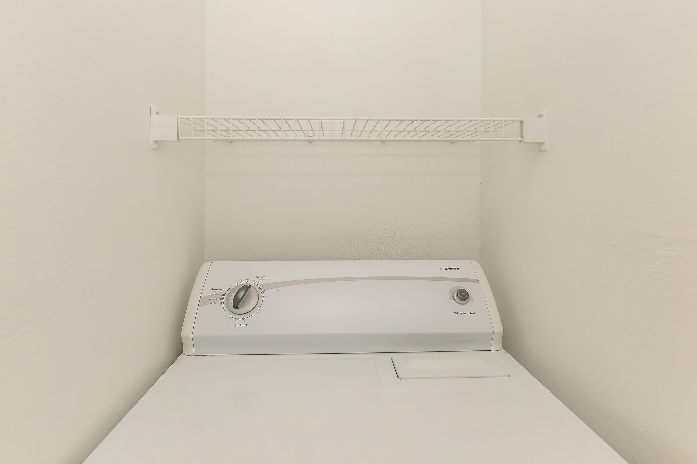 A top-loading washing machine situated in a small laundry area, with a metal wire shelf mounted above it against a plain wall. The machine features a control panel with dials for adjusting settings. The overall space is well-lit and has a minimalist design.