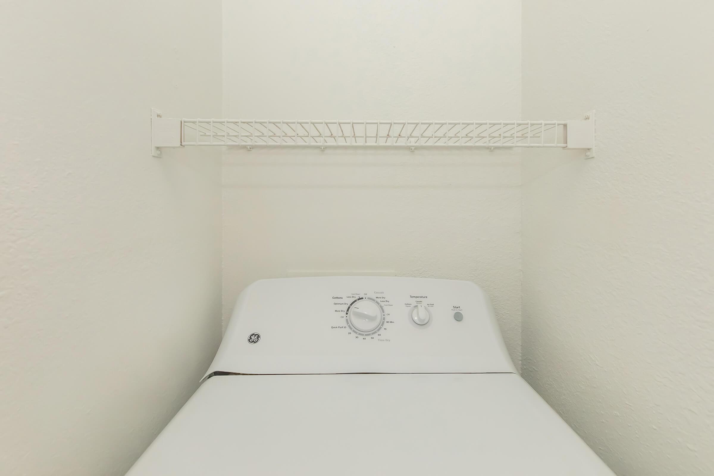 A white washing machine is positioned in a small, narrow laundry space with light-colored walls. Above the machine, there is a white wire shelf for storage. The overall setting is clean and minimalistic, emphasizing the compactness of the laundry area.