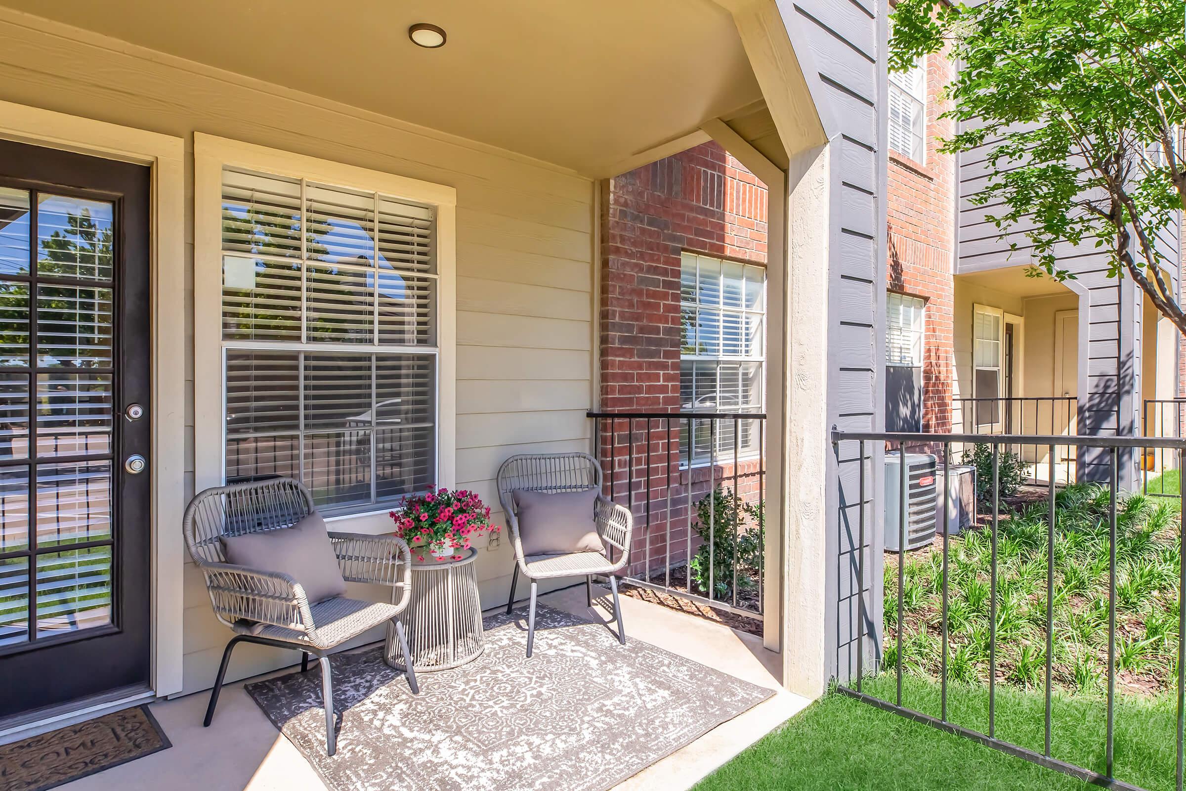 A welcoming apartment balcony featuring two stylish chairs, a small table adorned with a potted plant, and a decorative rug. The space is brightened by large windows and surrounded by greenery, creating a cozy outdoor seating area.