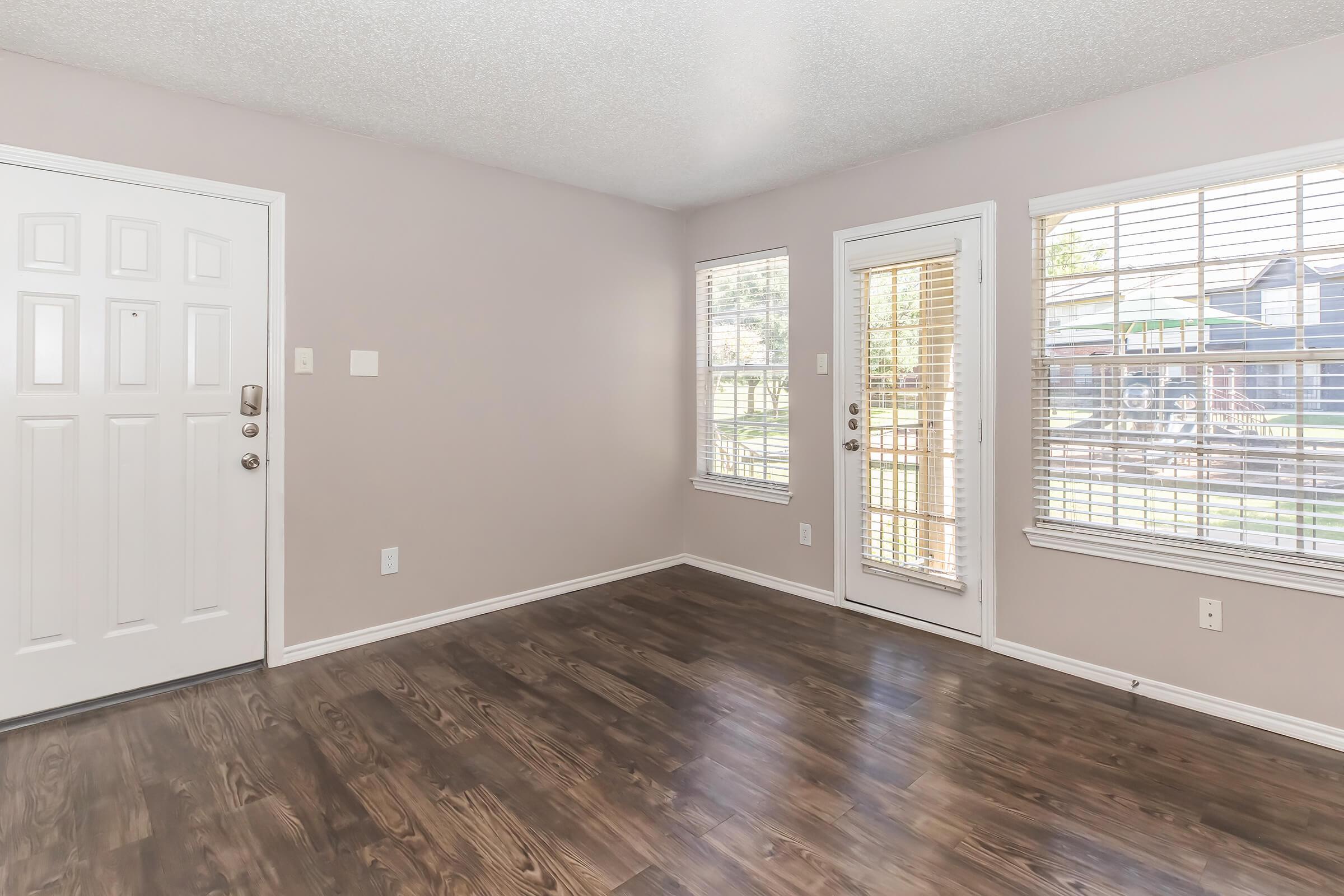 A well-lit, empty room featuring light-colored walls and hardwood flooring. There are two windows allowing natural light and a glass door leading outside. One wall has a front door with a simple design, and the space appears tidy and ready for furniture placement.