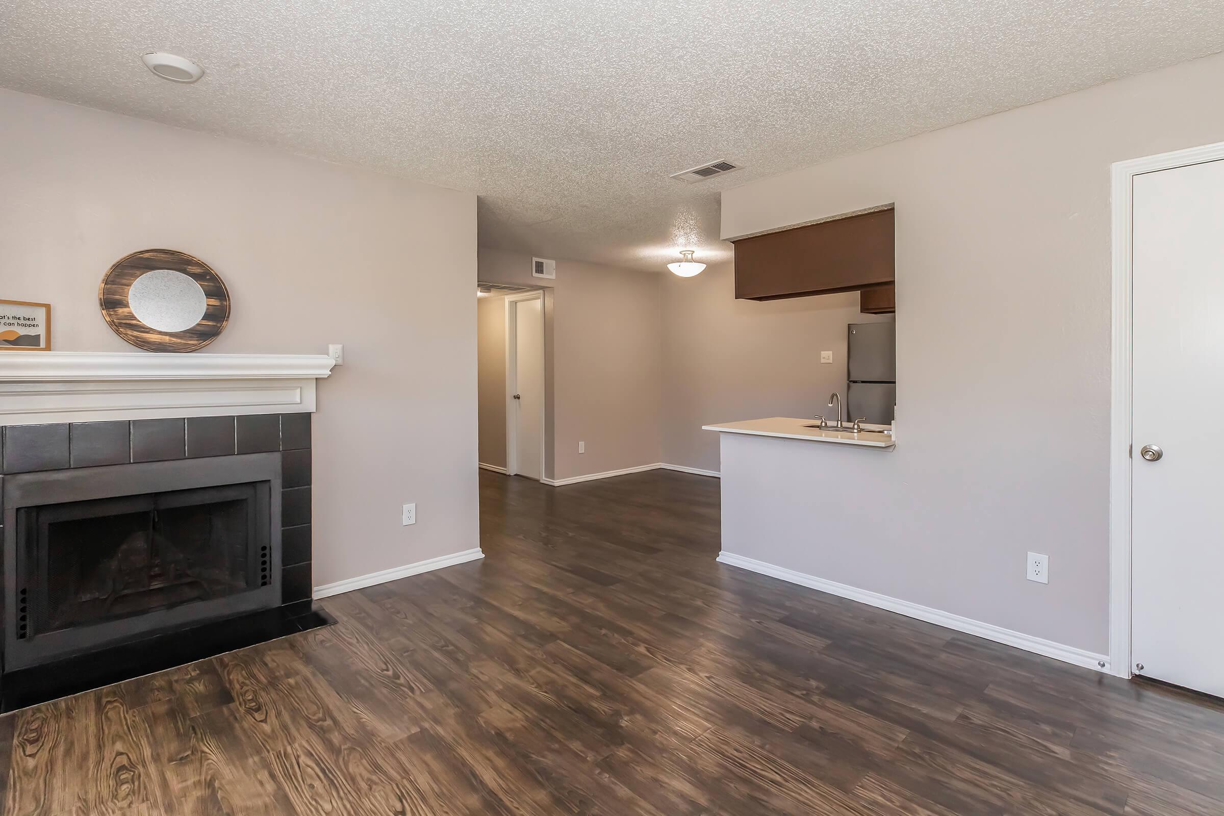 Spacious living area featuring a cozy fireplace, laminate flooring, and neutral-colored walls. A breakfast bar separates the kitchen, which includes stainless steel appliances. A hallway leads to additional rooms, with a door on the right providing access to other areas of the apartment. Bright, inviting atmosphere.