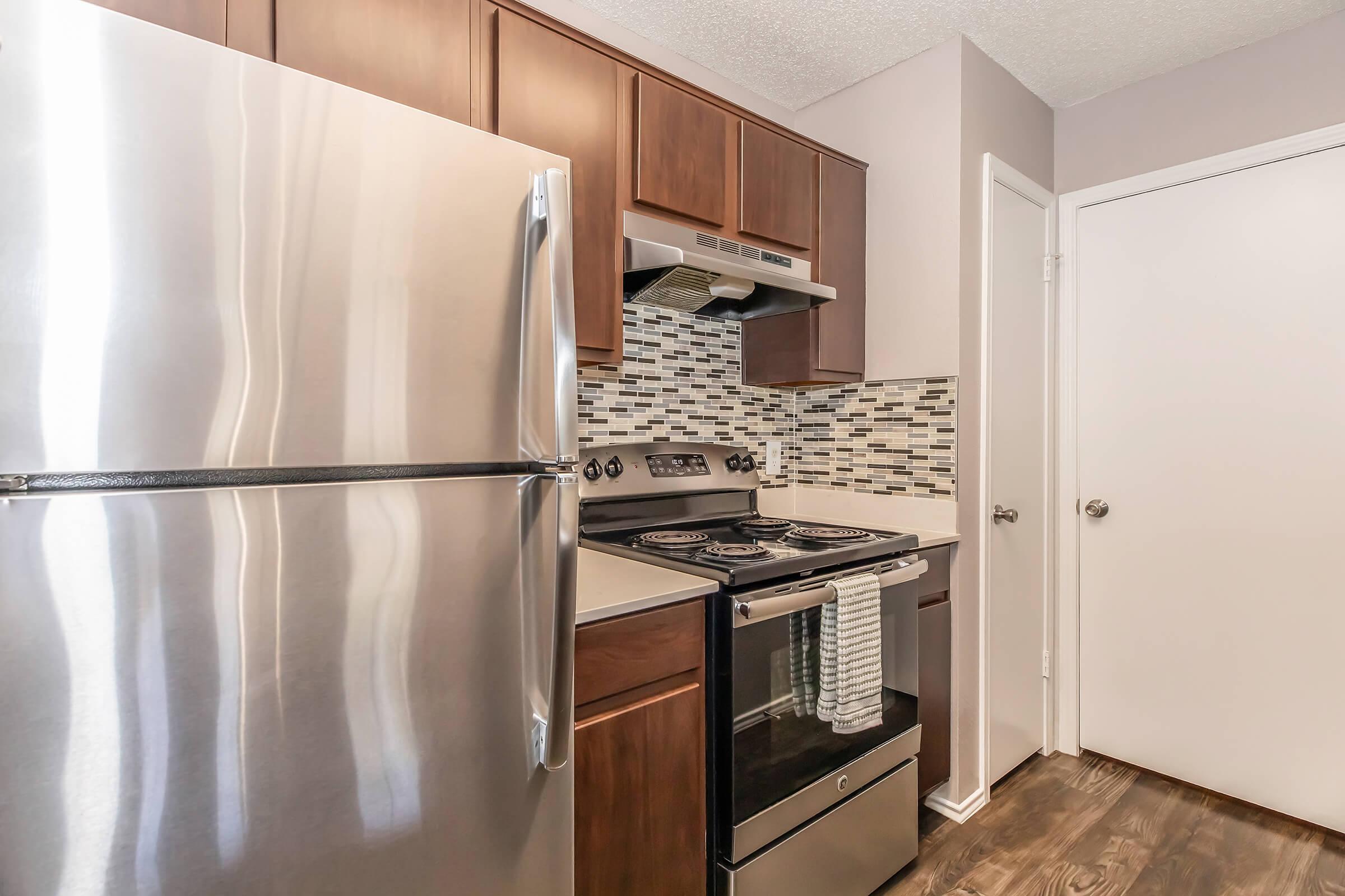 A modern kitchen featuring stainless steel appliances, including a refrigerator and an oven. The cabinets are dark brown, and the backsplash consists of small, multicolored tiles. The flooring is dark wood, and a white door is visible in the background.