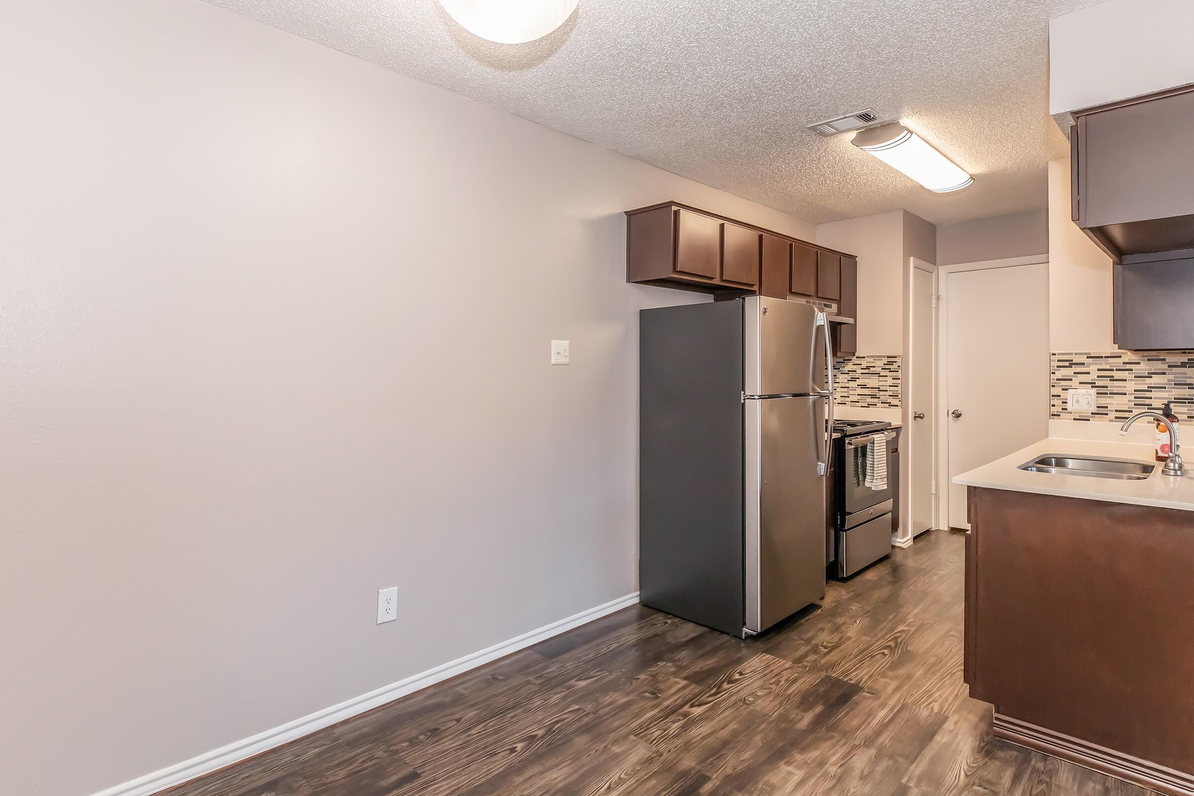 A modern kitchen featuring stainless steel appliances, including a refrigerator and stove. The cabinetry is dark wood, with a tiled backsplash. The flooring is dark laminate, and the walls are painted a light color. A small window and door lead to another area, creating an open feel.