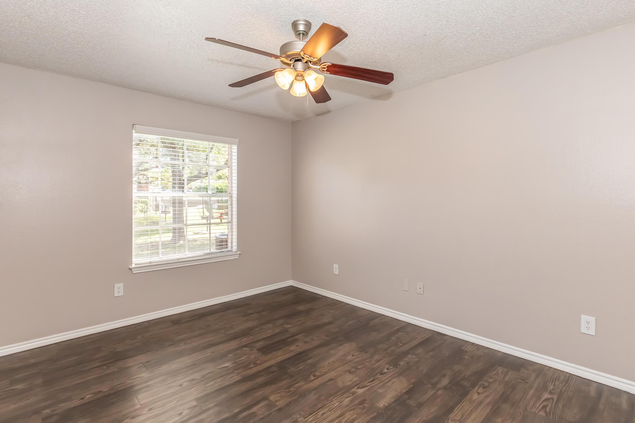 Empty room with light brown walls and a ceiling fan featuring four lights. A window allows natural light, revealing greenery outside. The floor is dark hardwood, and there are no furnishings or decorations visible. The space appears clean and ready for occupancy.