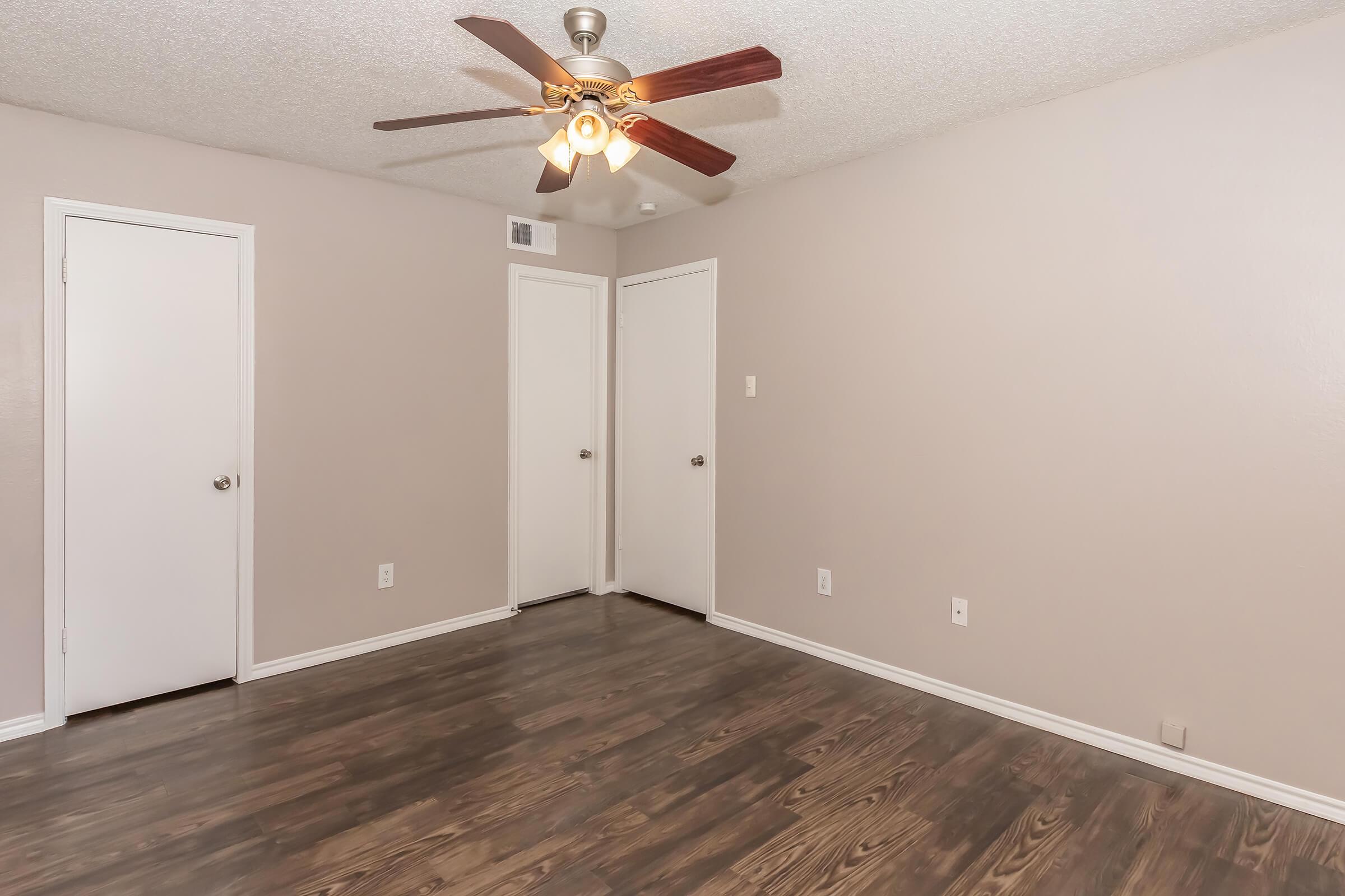 Interior of a room featuring a ceiling fan with five blades, light bulb, and textured walls painted in a neutral tone. The floor is dark wood laminate. There are two closed doors on the right side, and the room appears empty and well-lit, giving a spacious feel.