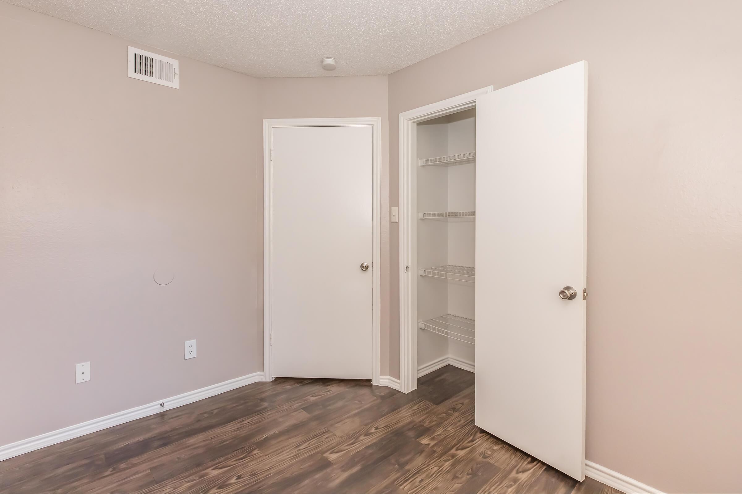 A small room with light beige walls and dark wood laminate flooring. There's a closed white door on the right leading to a closet with wire shelves. Another closed door is visible at the back wall, and a vent is located above the closet door. The room is well-lit and minimalistic.