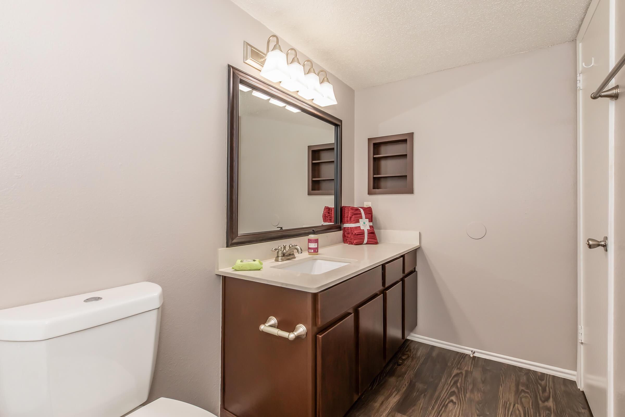 A well-lit bathroom featuring a white toilet and a dark wood vanity with a sink. Above the vanity, there is a large mirror and three light fixtures. The walls are painted a soft shade of beige, and there are shelves on the wall. A couple of red towels are neatly placed on the counter, adding a pop of color.
