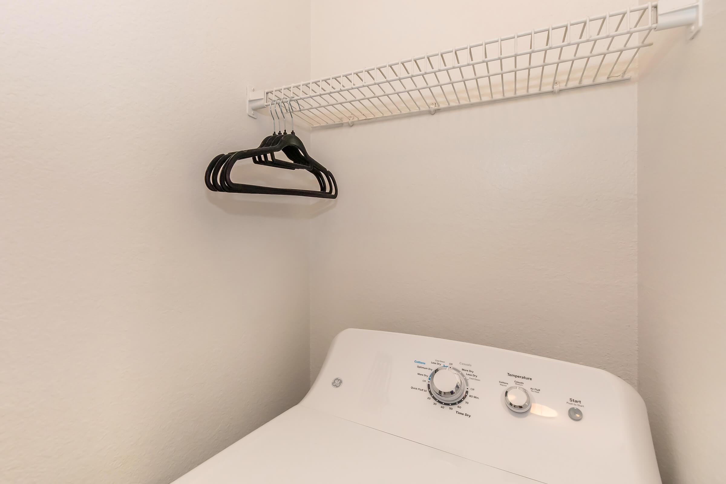 A laundry room corner featuring a white washing machine with a settings dial, and a shelf above it with five black hangers. The walls are plain and light-colored, providing a clean and minimalistic appearance.
