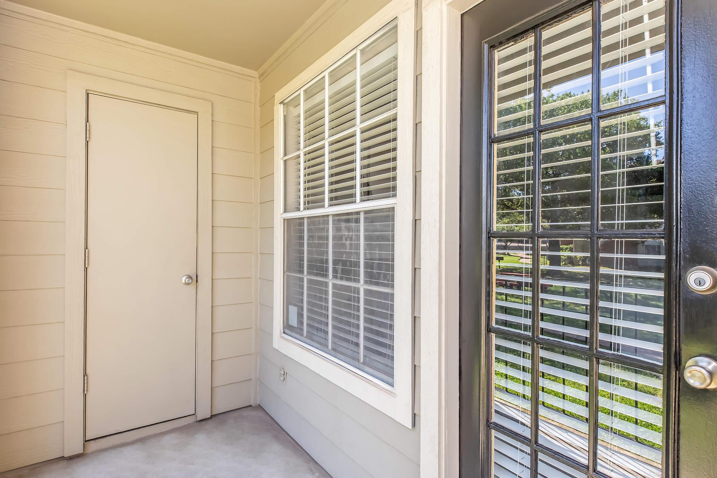 A view of an entrance featuring two doors, one solid and one with multiple panes of glass. There is a large window beside the door, with white blinds partially open, allowing for natural light and a view of greenery outside. The exterior is light-colored siding, creating a welcoming atmosphere.
