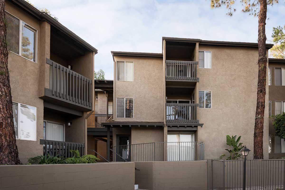 A view of a multi-unit apartment building with a tan exterior. The building features balconies and large windows, surrounded by trees. A gated entrance and decorative light fixtures are visible in the foreground, creating a welcoming atmosphere.