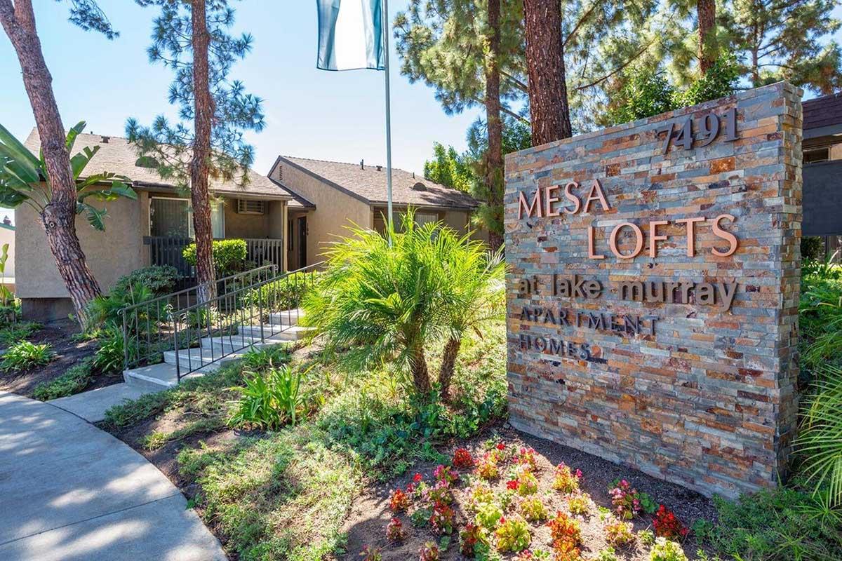 A landscaped entrance of Mesa Lofts at Lake Murray, featuring a stone sign with the name "Mesa Lofts" prominently displayed. Lush greenery and palm trees frame the pathway leading to the apartment complex, creating an inviting and peaceful atmosphere.