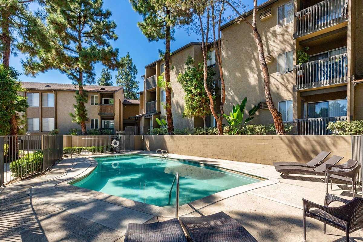 A serene pool area surrounded by greenery, featuring a rectangular swimming pool with steps, lounge chairs, and apartments in the background. Tall trees provide shade, creating a relaxing atmosphere in an inviting residential complex