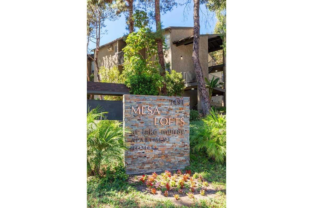 Sign for "Mesa Lofts" with a stone base, surrounded by greenery and colorful flowers. In the background, you can see part of the building with balconies and trees nearby. The setting appears to be a residential area with a warm, inviting atmosphere.
