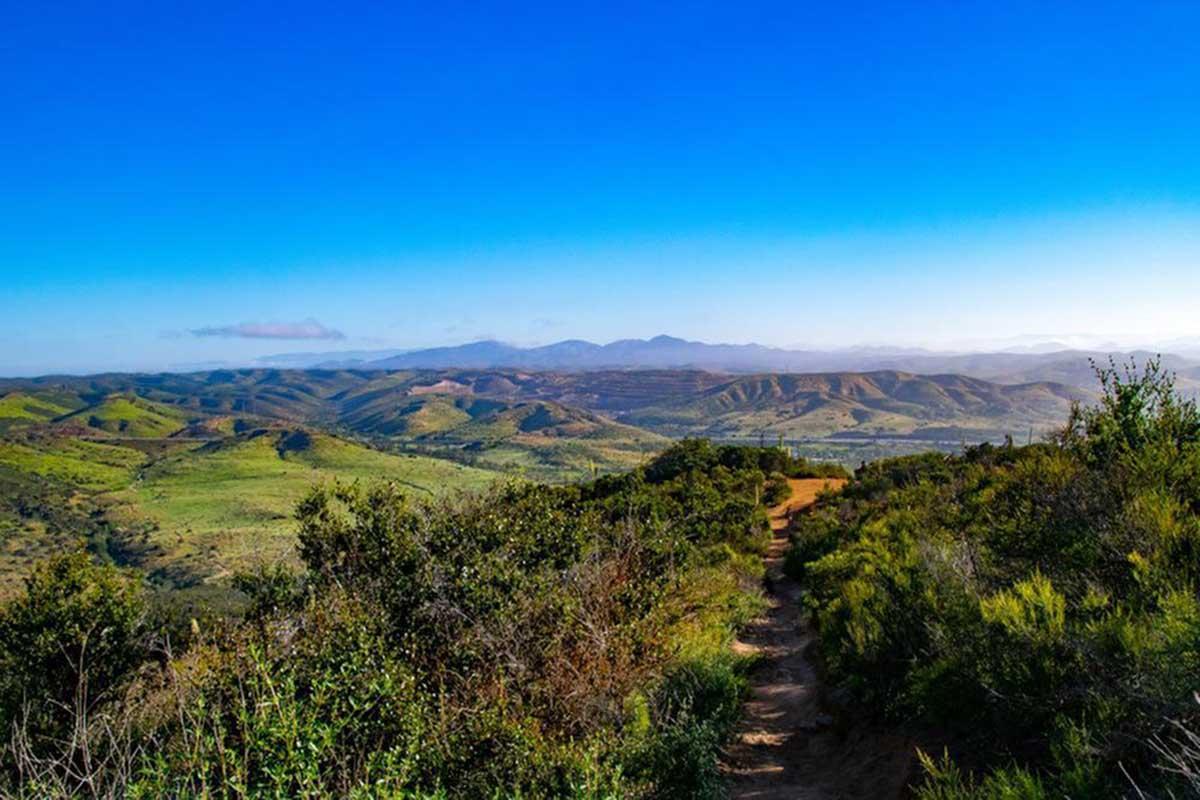 A scenic view of rolling green hills under a clear blue sky, with a dirt pathway winding through the landscape. In the distance, majestic mountains can be seen, creating a picturesque and tranquil natural setting. The scene captures the beauty of the outdoors and promotes a sense of peace and adventure.
