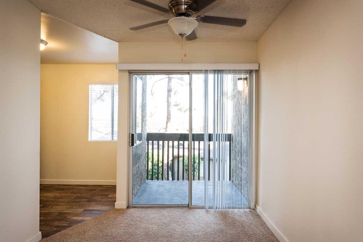 Interior view of a room featuring a sliding glass door that opens to a balcony, with a ceiling fan above. Natural light enters through a nearby window, illuminating the carpeted floor and adjacent walls. Modern and minimalistic design elements are prominent in the space.