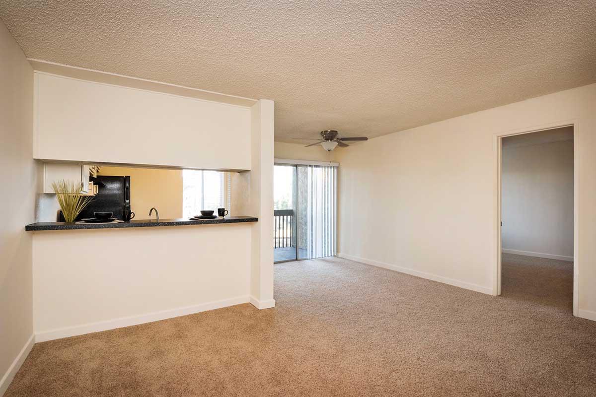 A spacious living area featuring a small kitchenette with a bar counter, light-colored walls, and beige carpeting. A sliding glass door leads to a balcony, and there’s a ceiling fan for ventilation. The room is bright and inviting, with a neutral color palette and minimalistic decor.