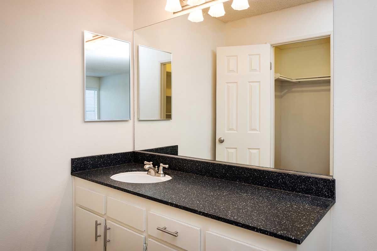 A modern bathroom with a dark speckled countertop, a white sink, and two mirrors above it. The walls are painted a light color, and a door leads to a closet. Soft lighting comes from a fixture with four bulbs above the mirrors. The overall design is clean and minimalist.