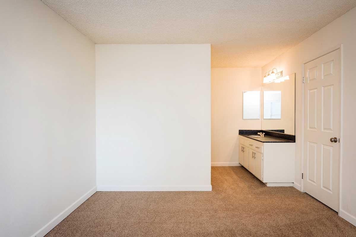 Empty room with beige carpet flooring and white walls. On one side, there is a small vanity area with a dark countertop and a mirror, providing a simple and clean space. A closed door is visible, leading to another area. Natural light fills the room, creating a bright and inviting atmosphere.